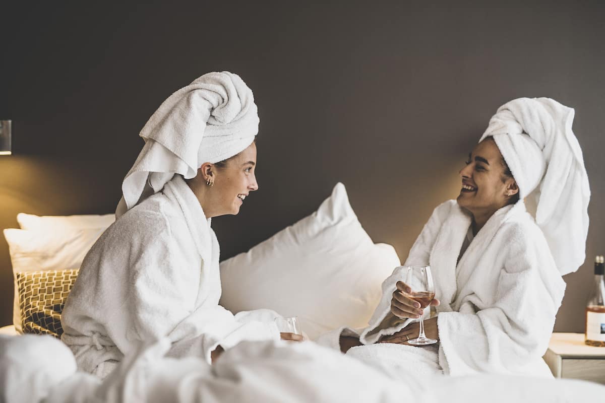 Two people in white bathrobes sitting on a bed with a bottle of wine and glasses.