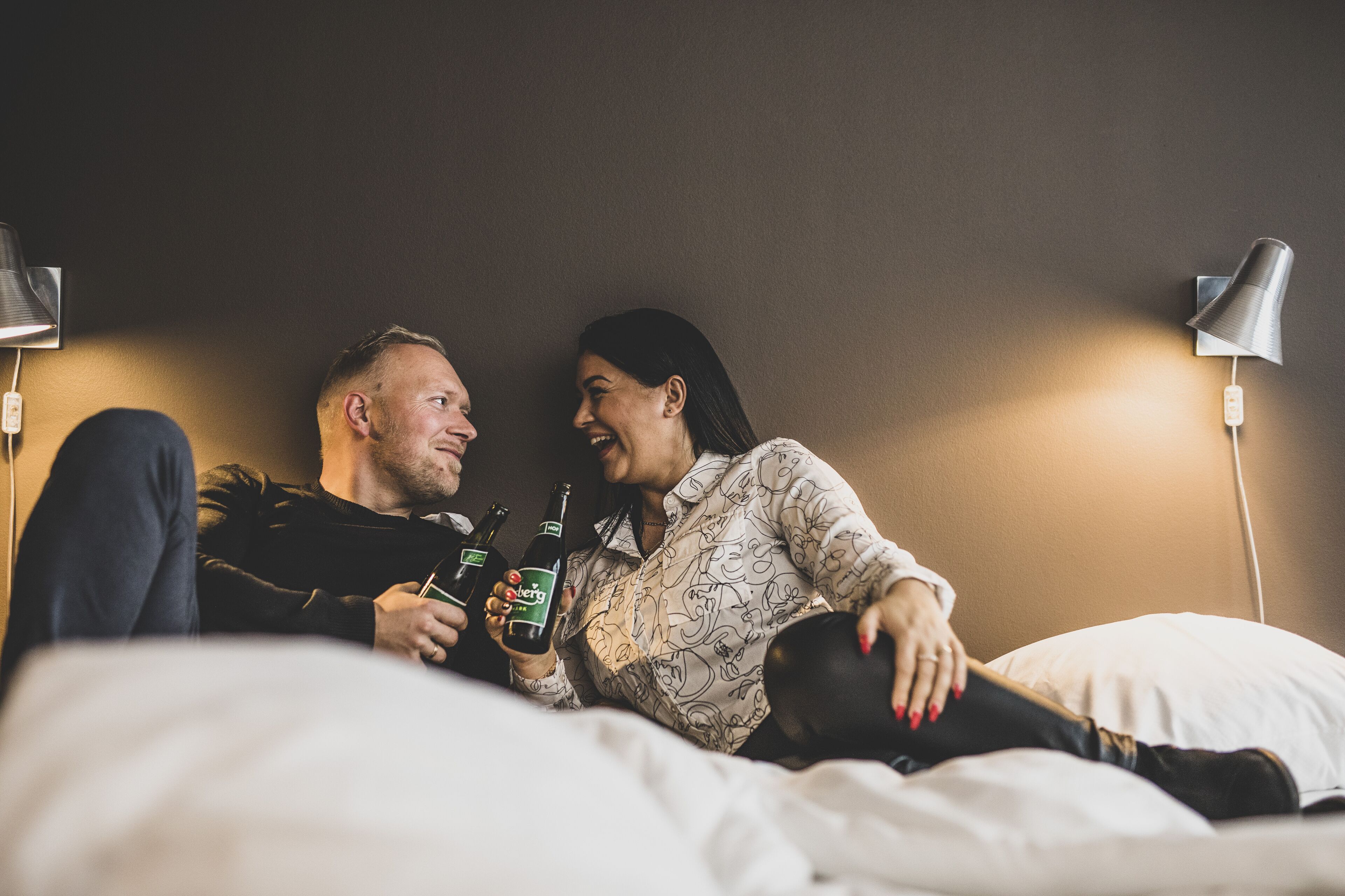 Um casal sentado na cama, com uma luminária e uma garrafa de cerveja.