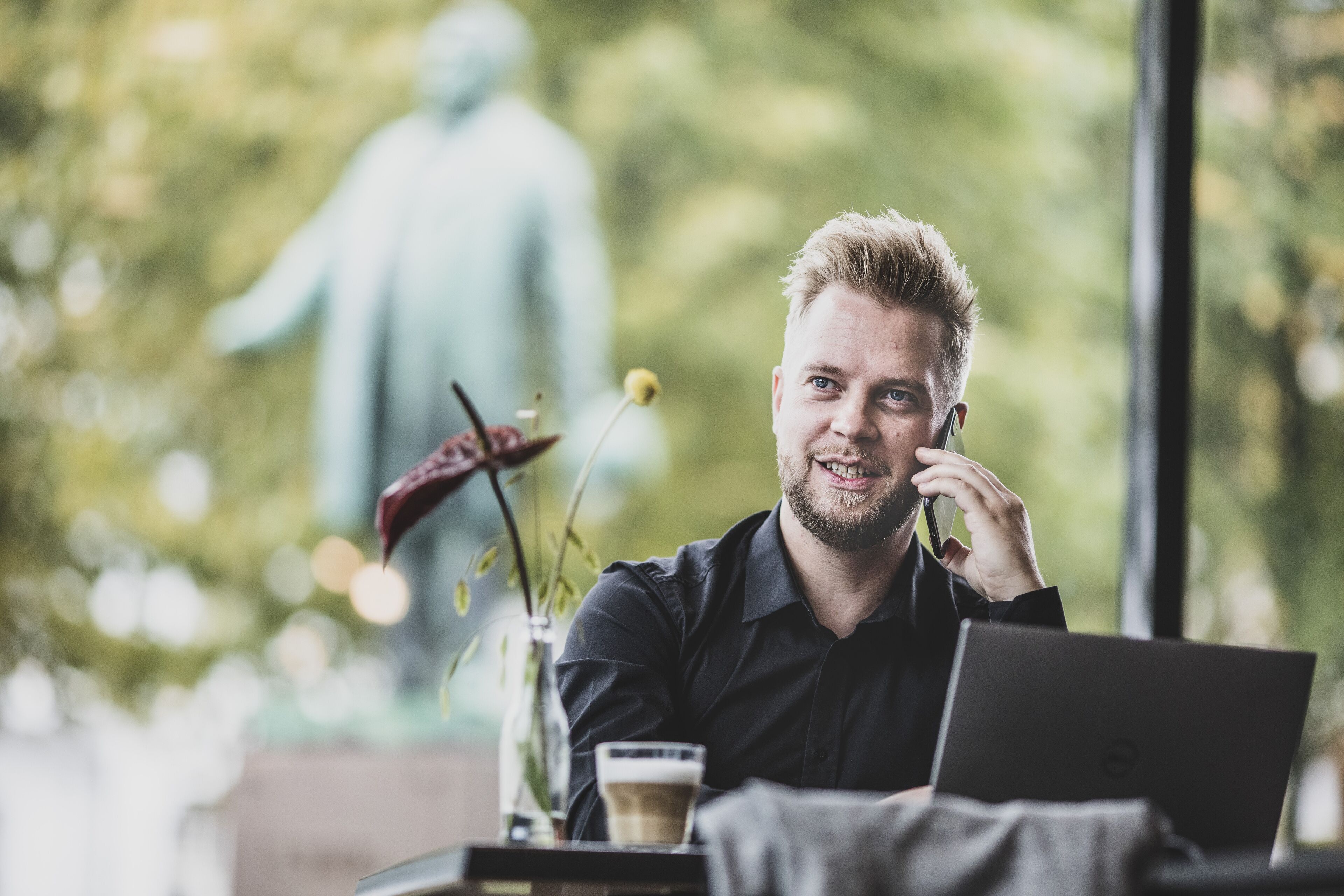 A person sitting at a table with a laptop and a cup, in a room with a statue in the background.