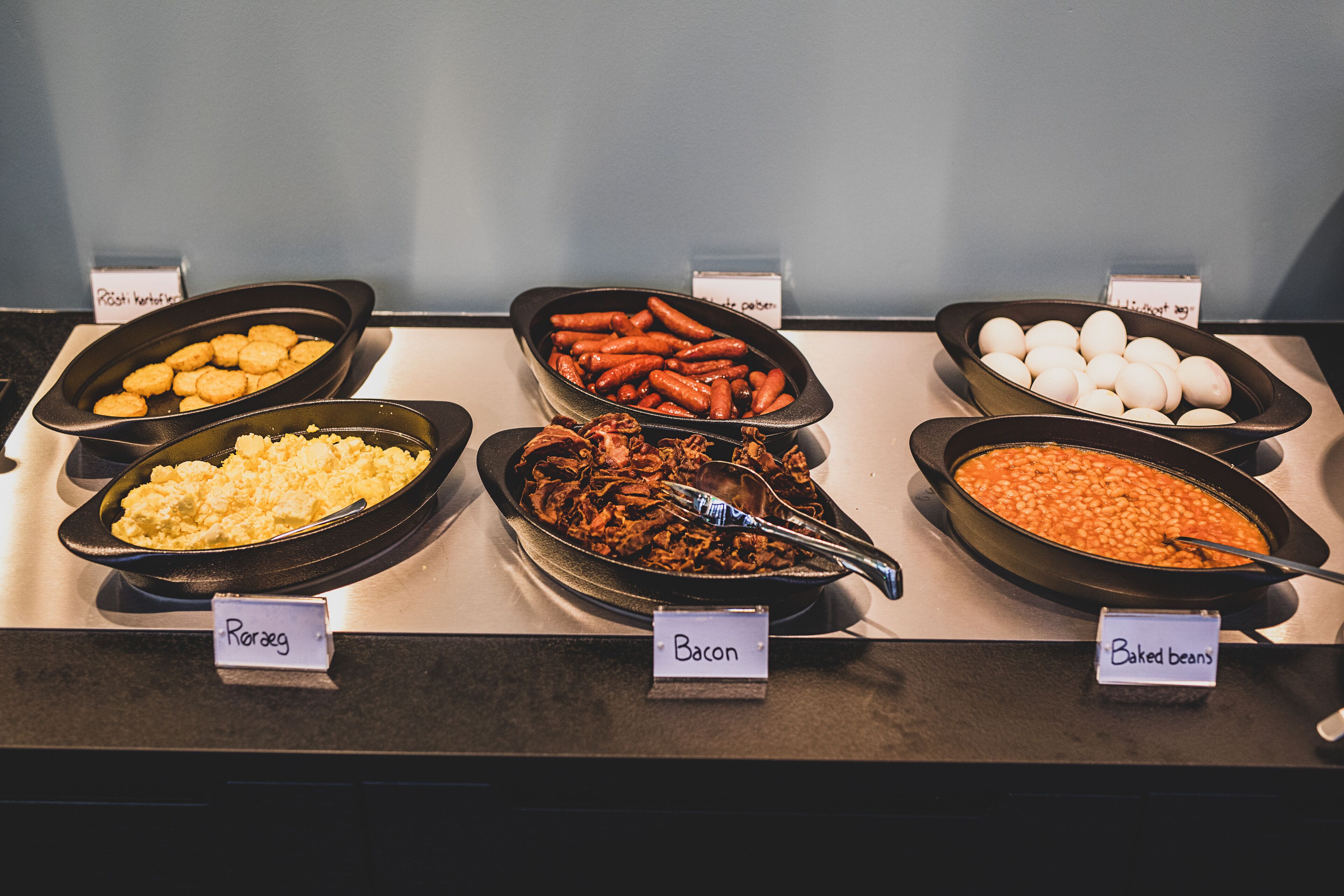 A buffet with labeled dishes including Röseg, Bacon, and Baked Beans.