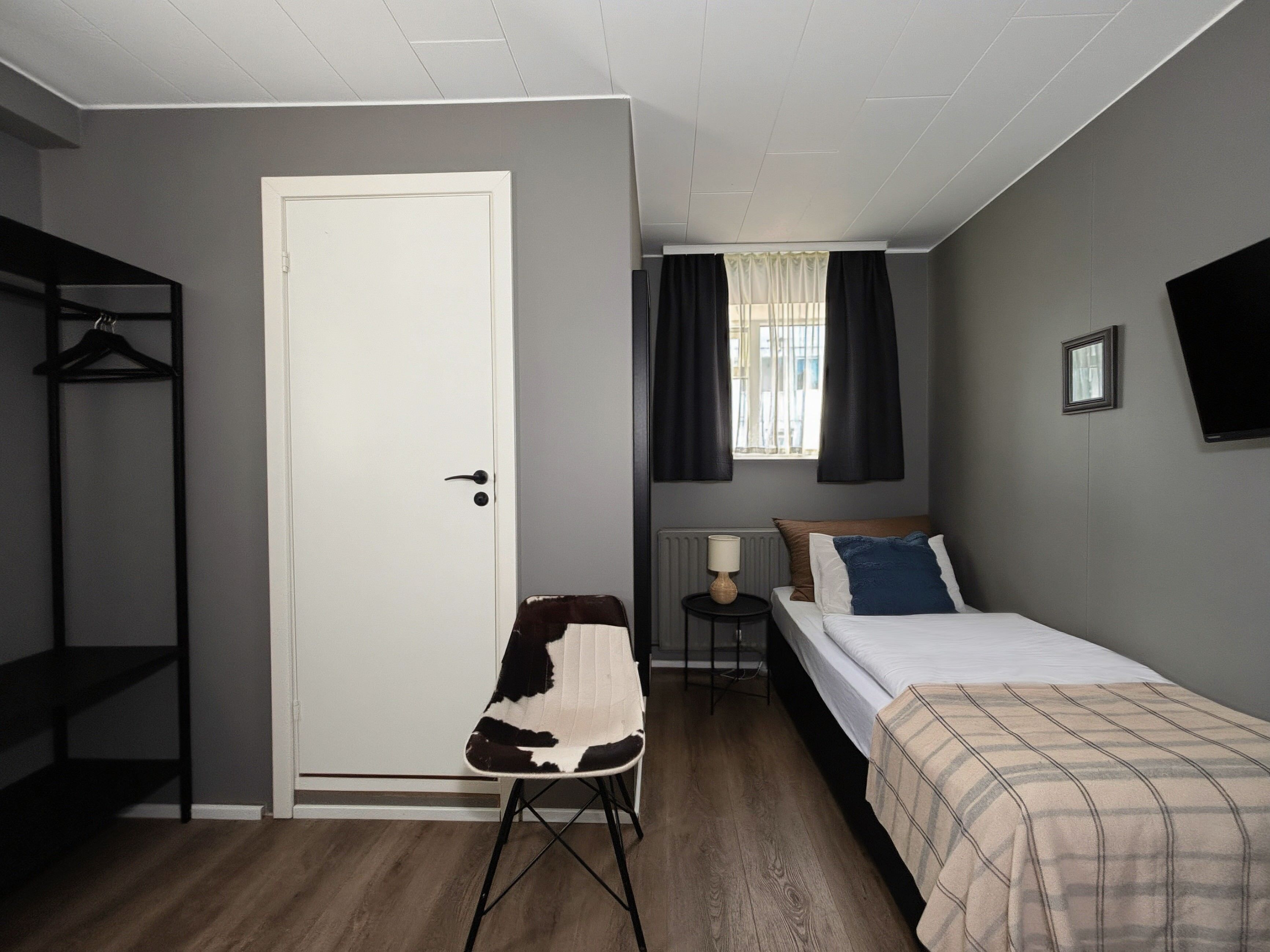 A bedroom with a bed, a chair, a radiator, a window with curtains, and a door.