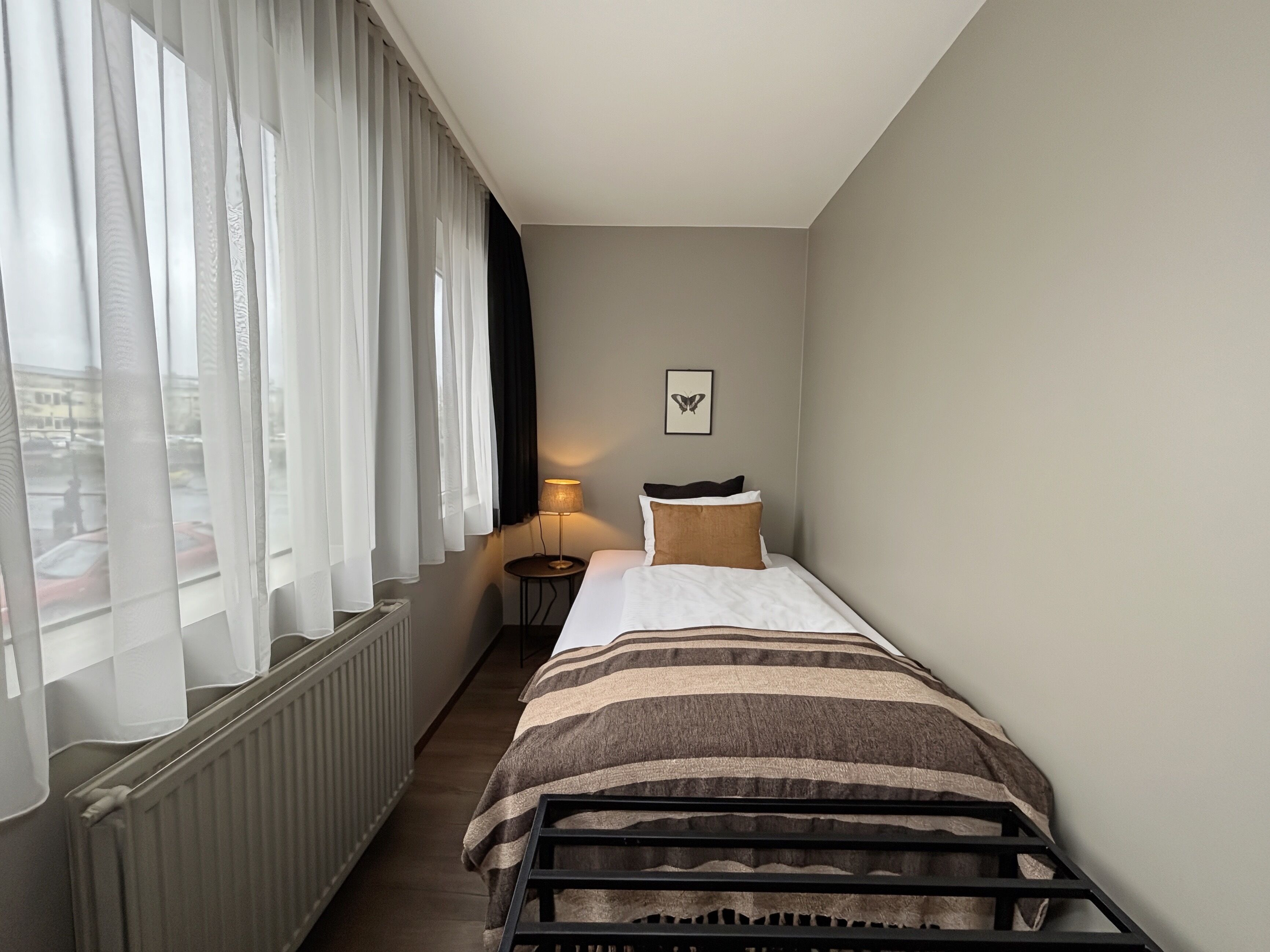 Habitación de hotel con cama, mesita de noche con lámpara, cuadro enmarcado en la pared y ventana con cortinas traslúcidas.