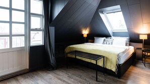 Ein Schlafzimmer mit schrägen Wänden, einem Bett mit gelber Tagesdecke, zwei Nachttischlampen und einem Fenster mit Blick ins Freie.