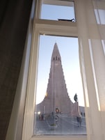 Vista a través de una ventana de un edificio alto y con techo puntiagudo, con una cruz en la parte superior, probablemente una iglesia o catedral, con gente caminando al frente.