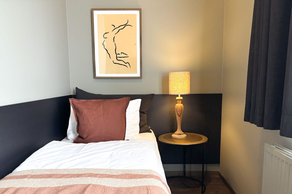 Une chambre d’hôtel avec un lit, une table de chevet, une lampe, un tableau encadré et une fenêtre avec des rideaux.