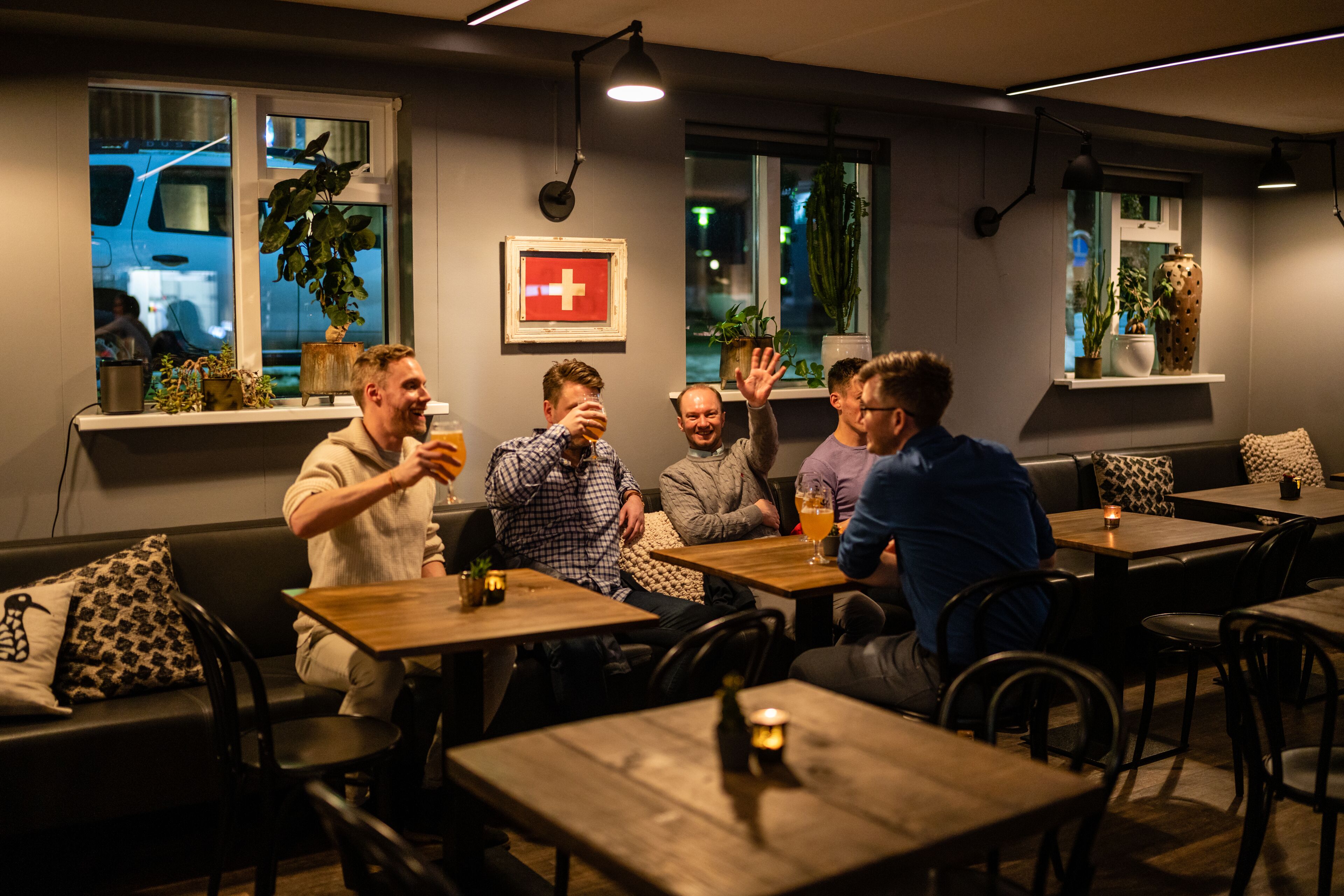 A group of people sitting at tables in a restaurant with wooden furniture, potted plants, and a Swiss flag on the wall.