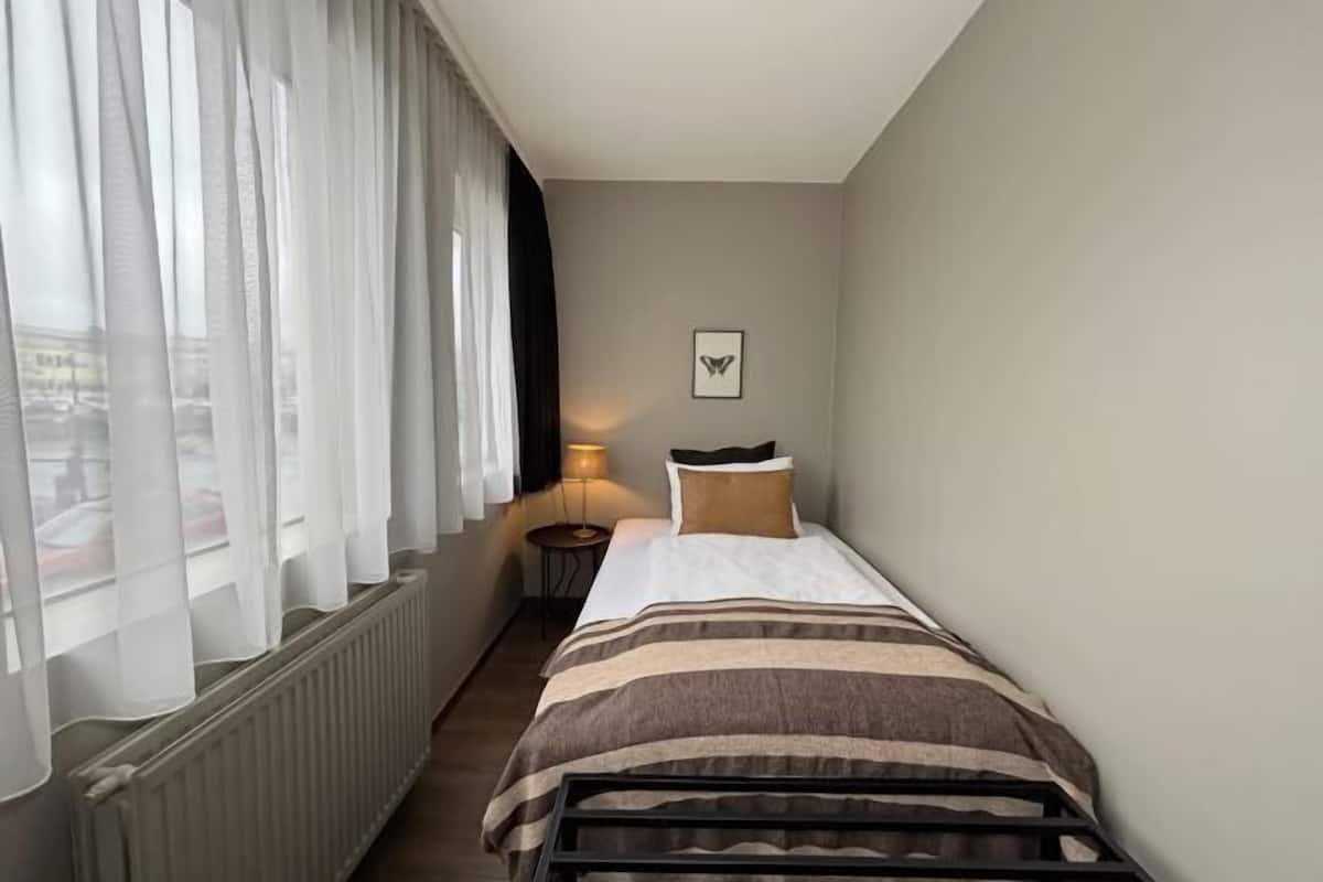 Ein Hotelzimmer mit einem Bett, einem Nachttisch, einer Lampe, einem Fenster mit durchsichtigen Vorhängen und einem gerahmten Bild an der Wand.