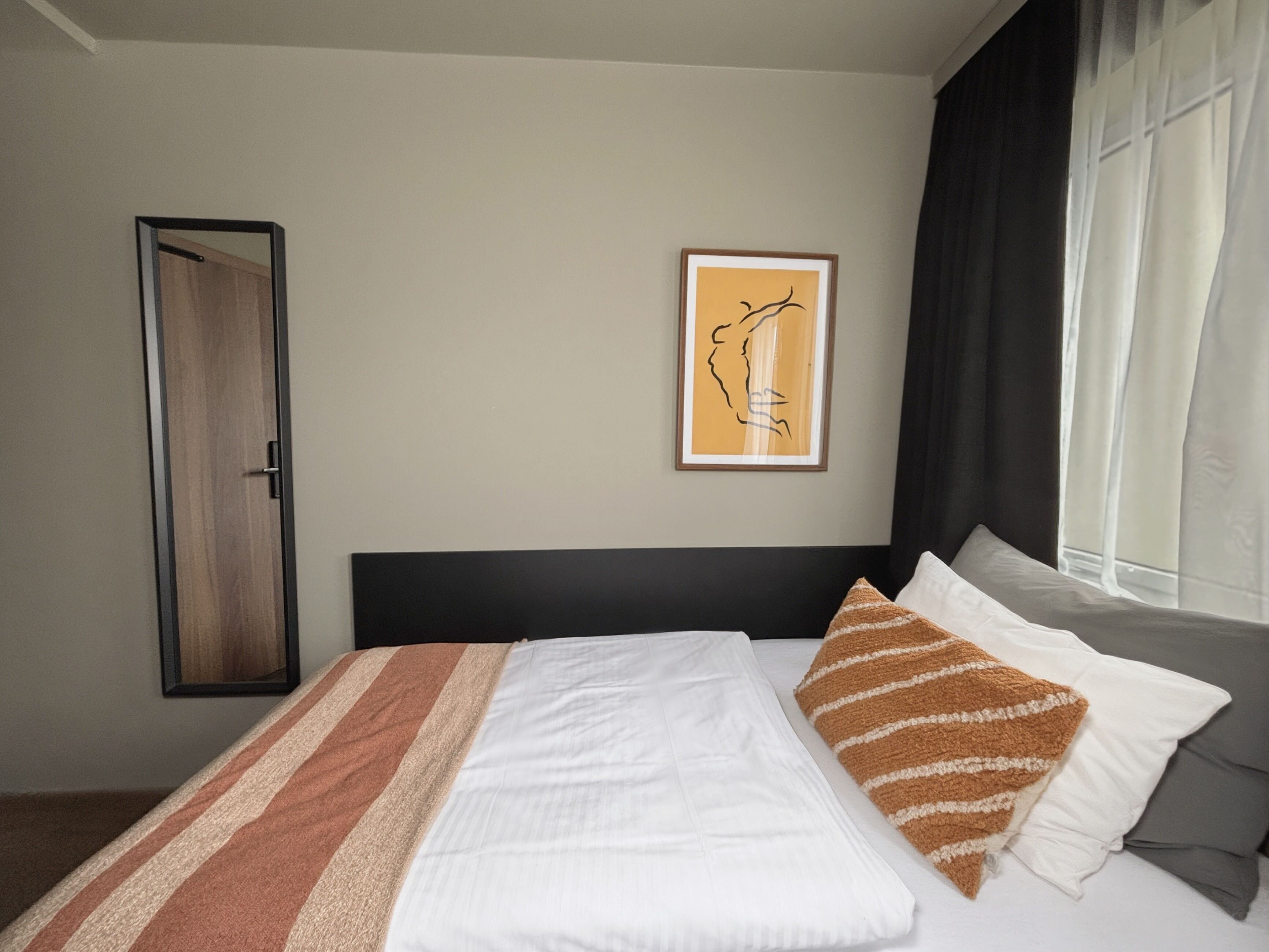 Habitación de hotel con cama, una obra de arte enmarcada en la pared, una puerta y una ventana con cortinas.