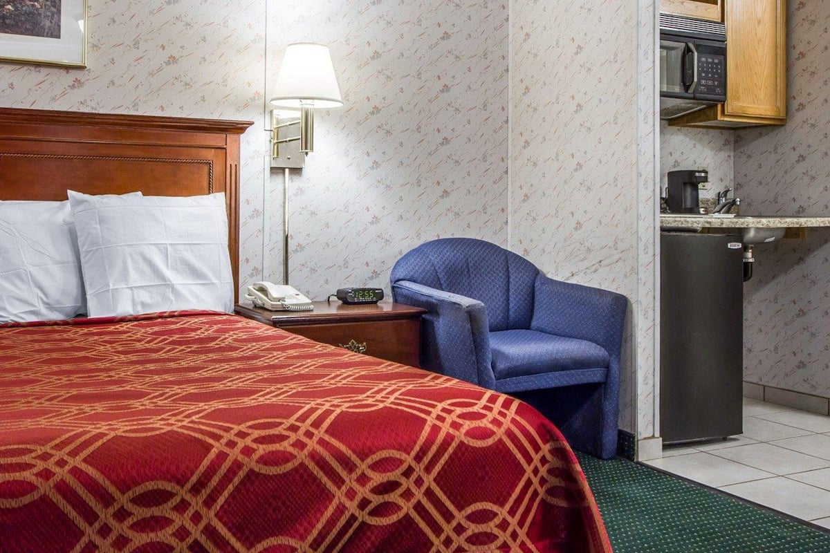 A hotel room with a bed, a chair, a desk with a lamp, a phone, a clock, and a kitchenette.