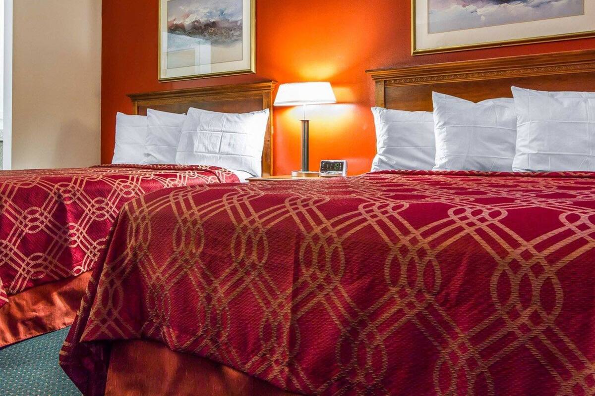 A hotel room with two beds, red patterned bedspreads, a nightstand with a lamp, and two framed pictures on the wall.