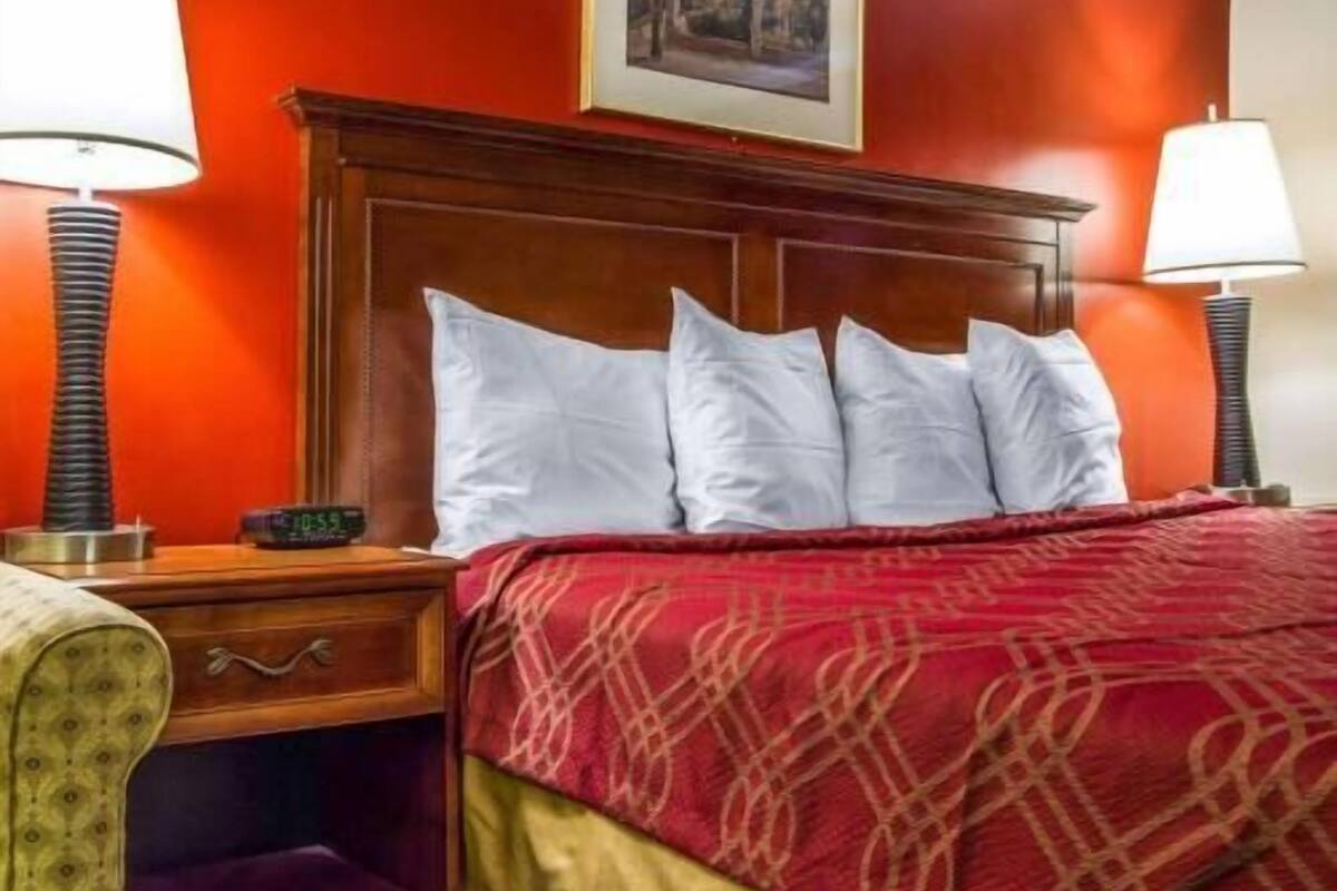 A hotel room with a wooden headboard, a red patterned bedspread, two bedside lamps, a nightstand with a clock, and a framed picture on the wall.