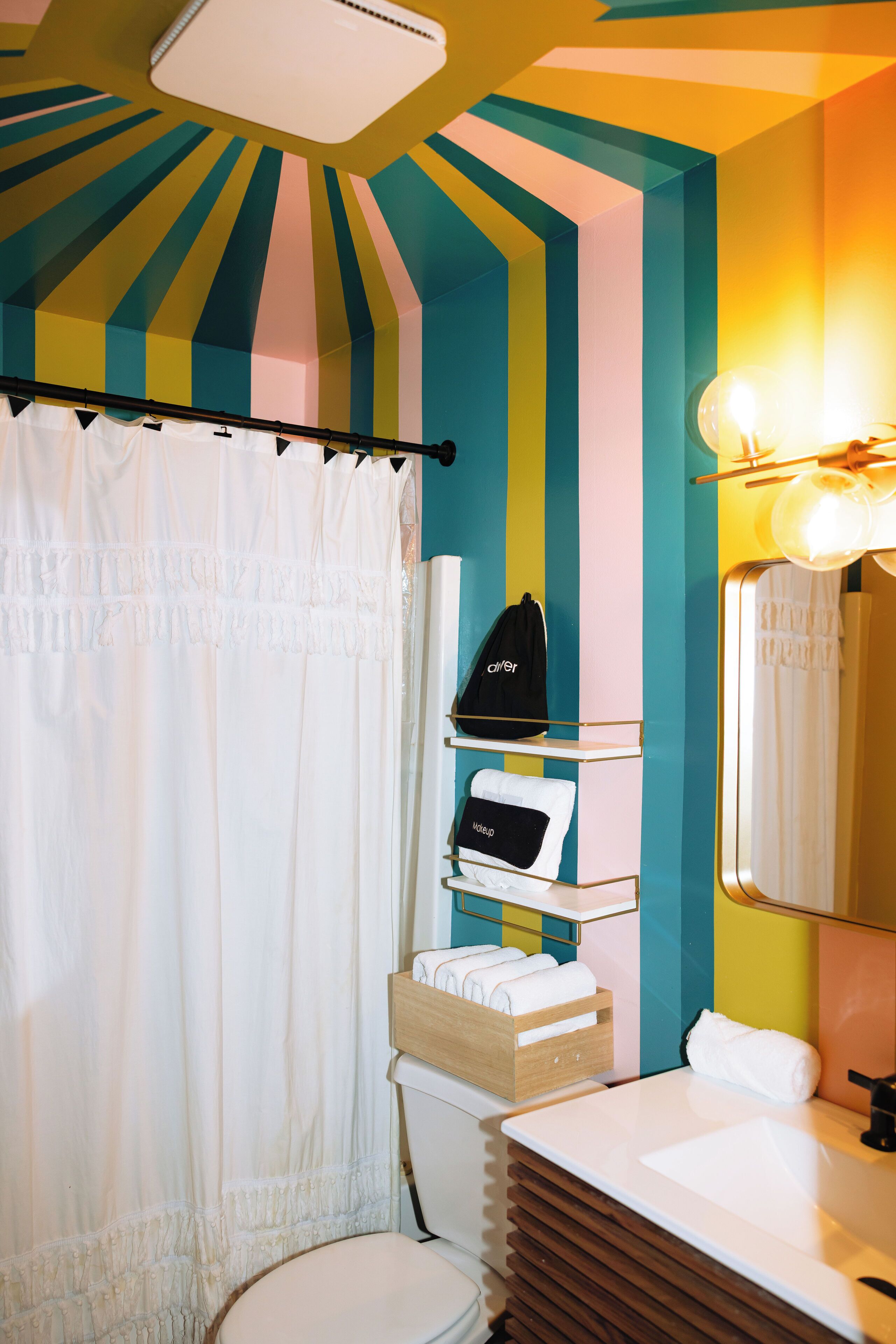 A bathroom with a striped wall design, a white toilet, a vanity with a mirror, and a shelf with a black bag and a makeup pouch.