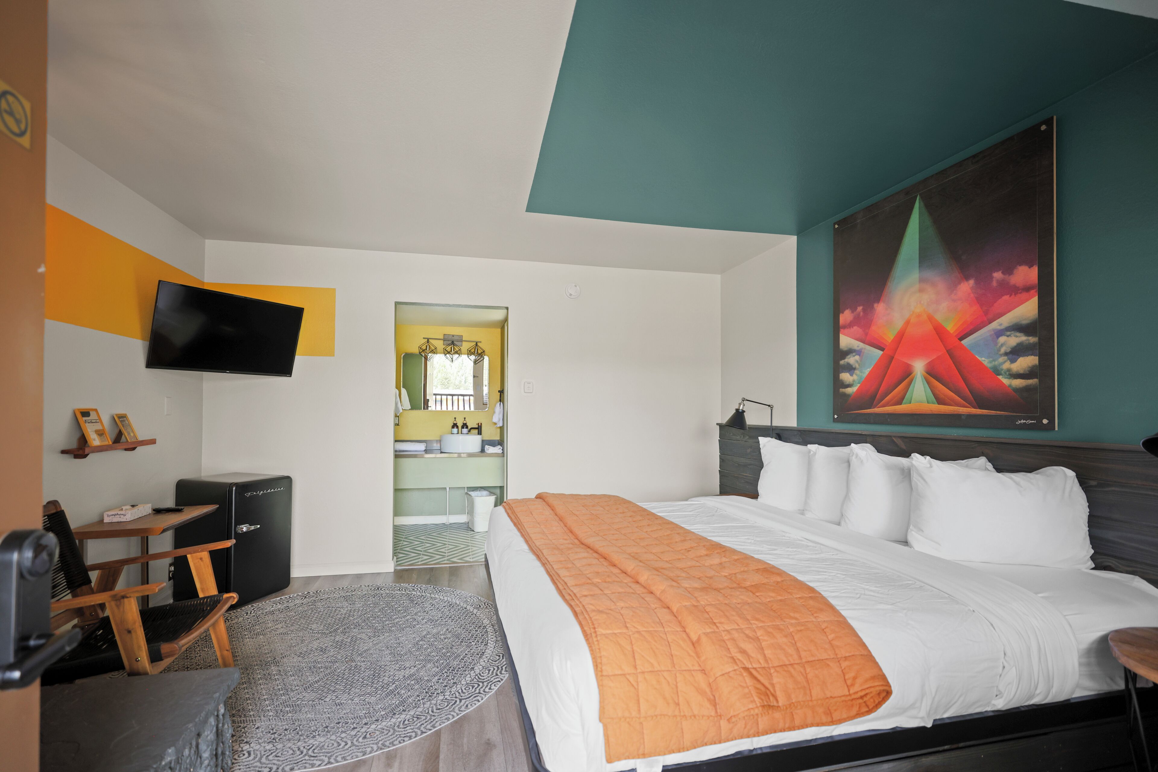 A modern hotel room with a large bed, a desk with a computer, a TV mounted on the wall, and a painting above the bed.