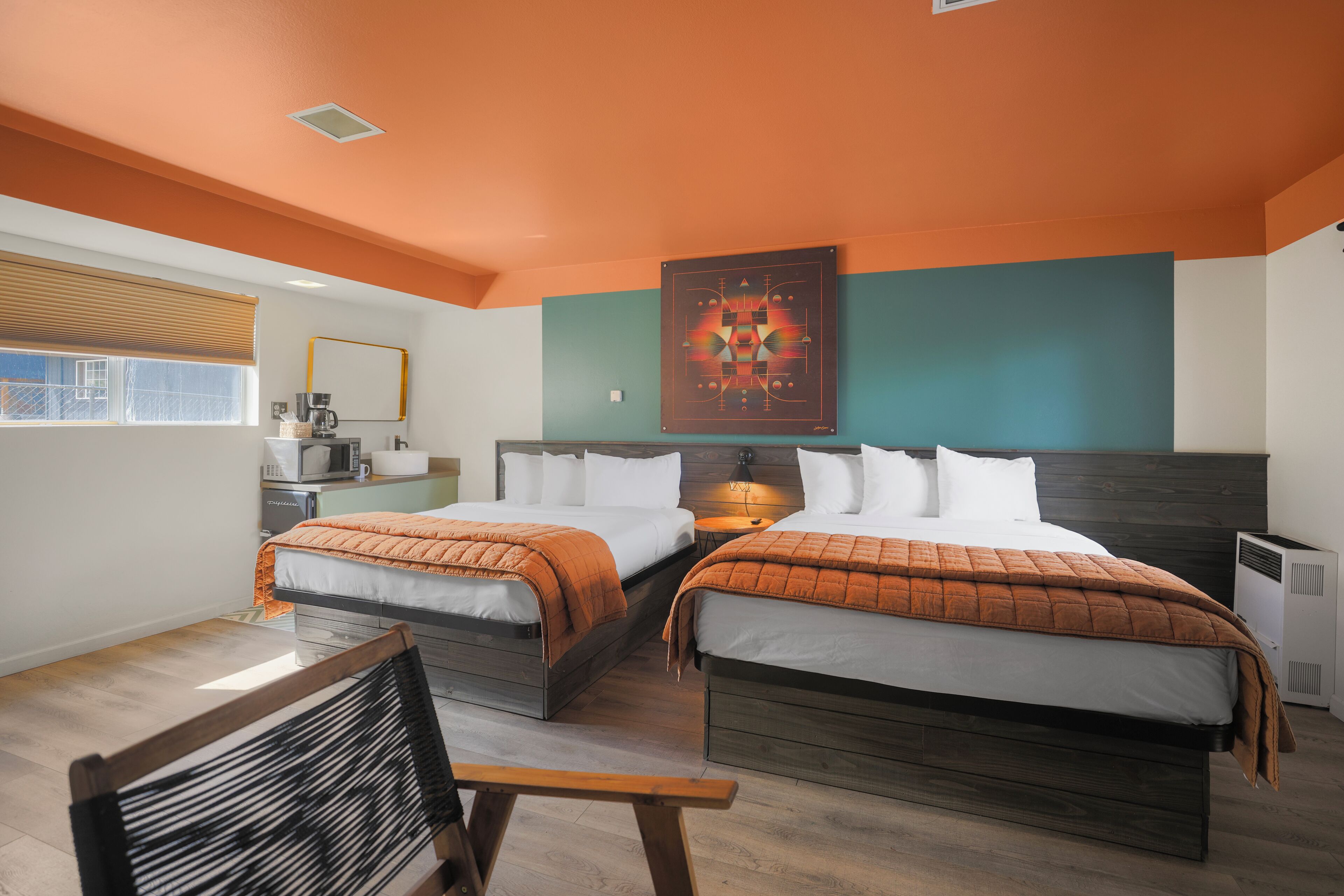 A hotel room with two beds, a wooden bench, a desk with a chair, a window with blinds, and a wall-mounted artwork.
