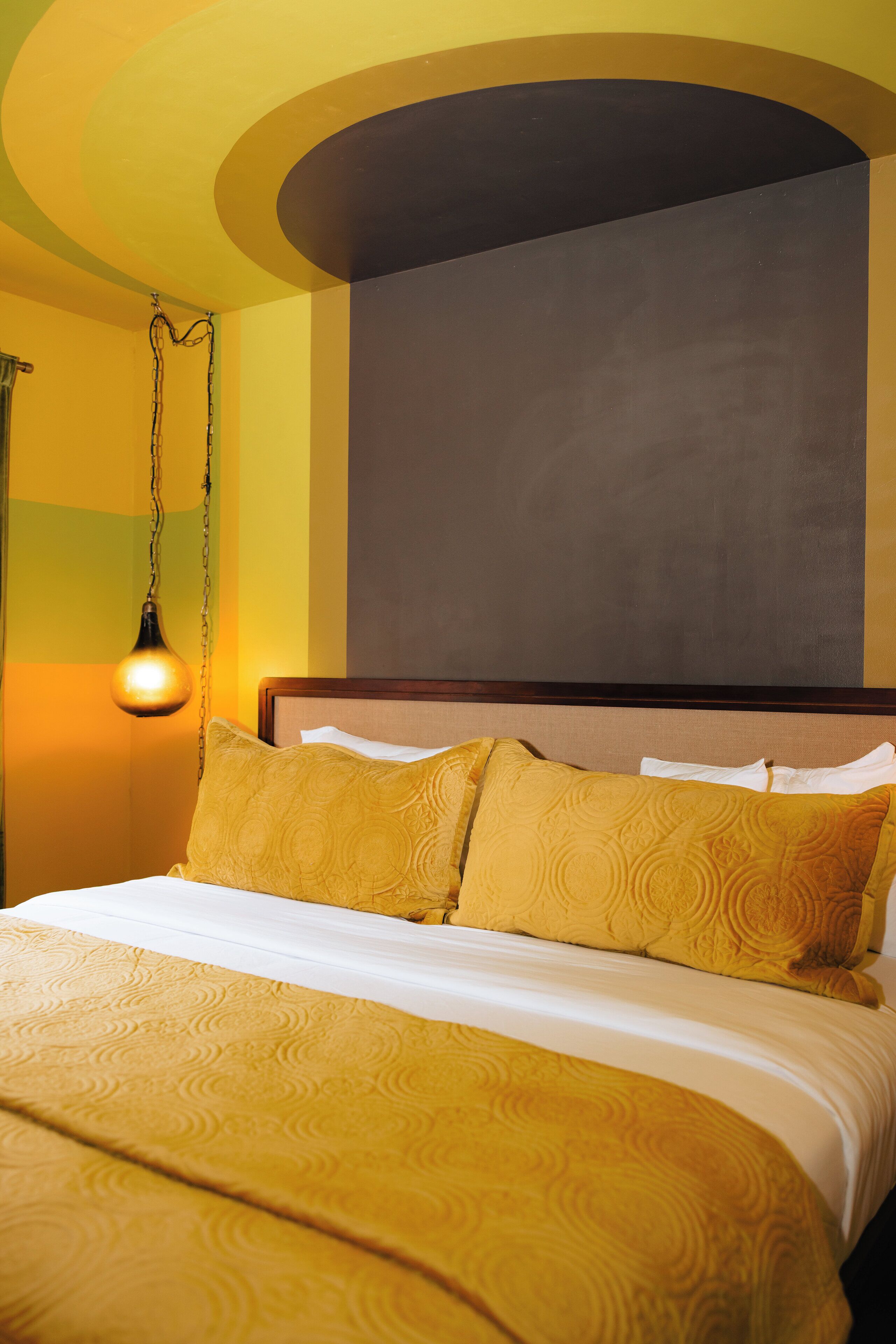 A hotel room with a bed, a hanging light fixture, and a plain wall.