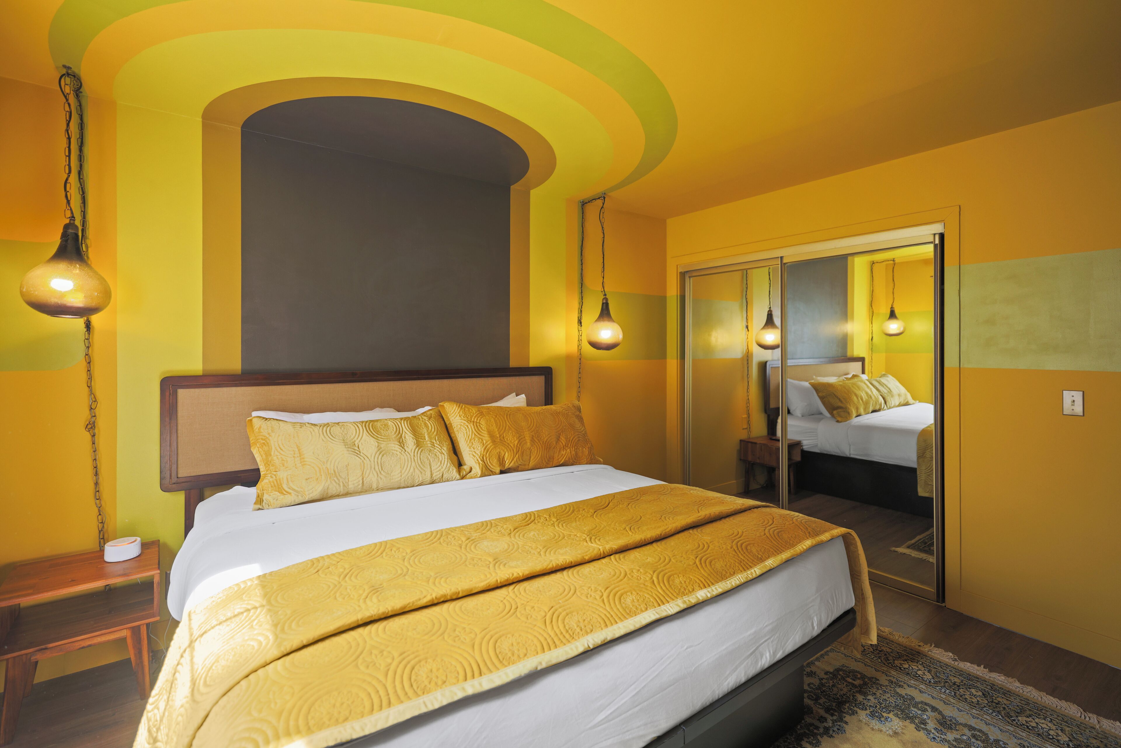 Habitación de hotel con una cama grande, una mesita de noche, un espejo y una puerta que da a otra habitación.