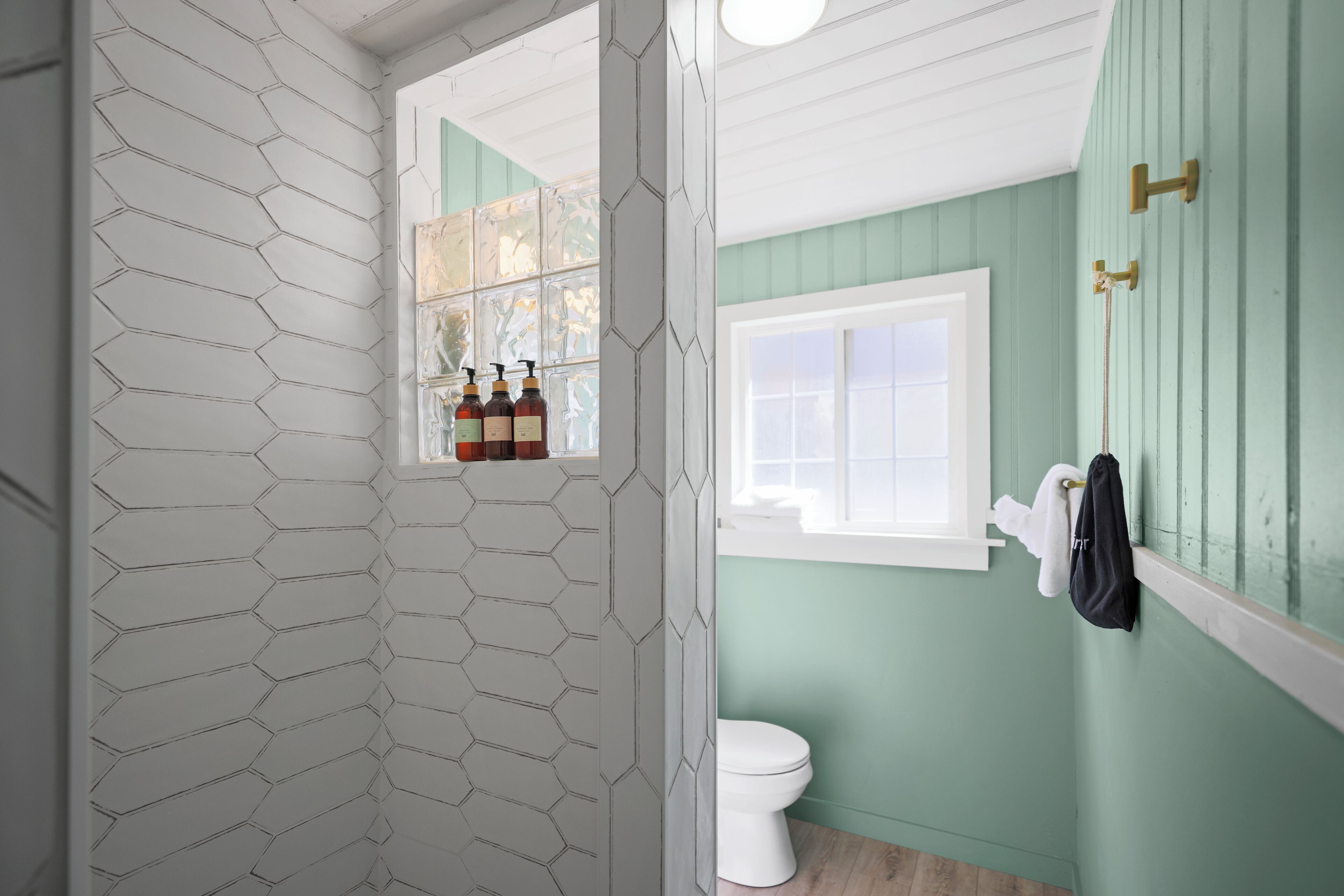 A bathroom with a white hexagonal tile wall, a green wall, a white toilet, and a window with bottles on the sill.