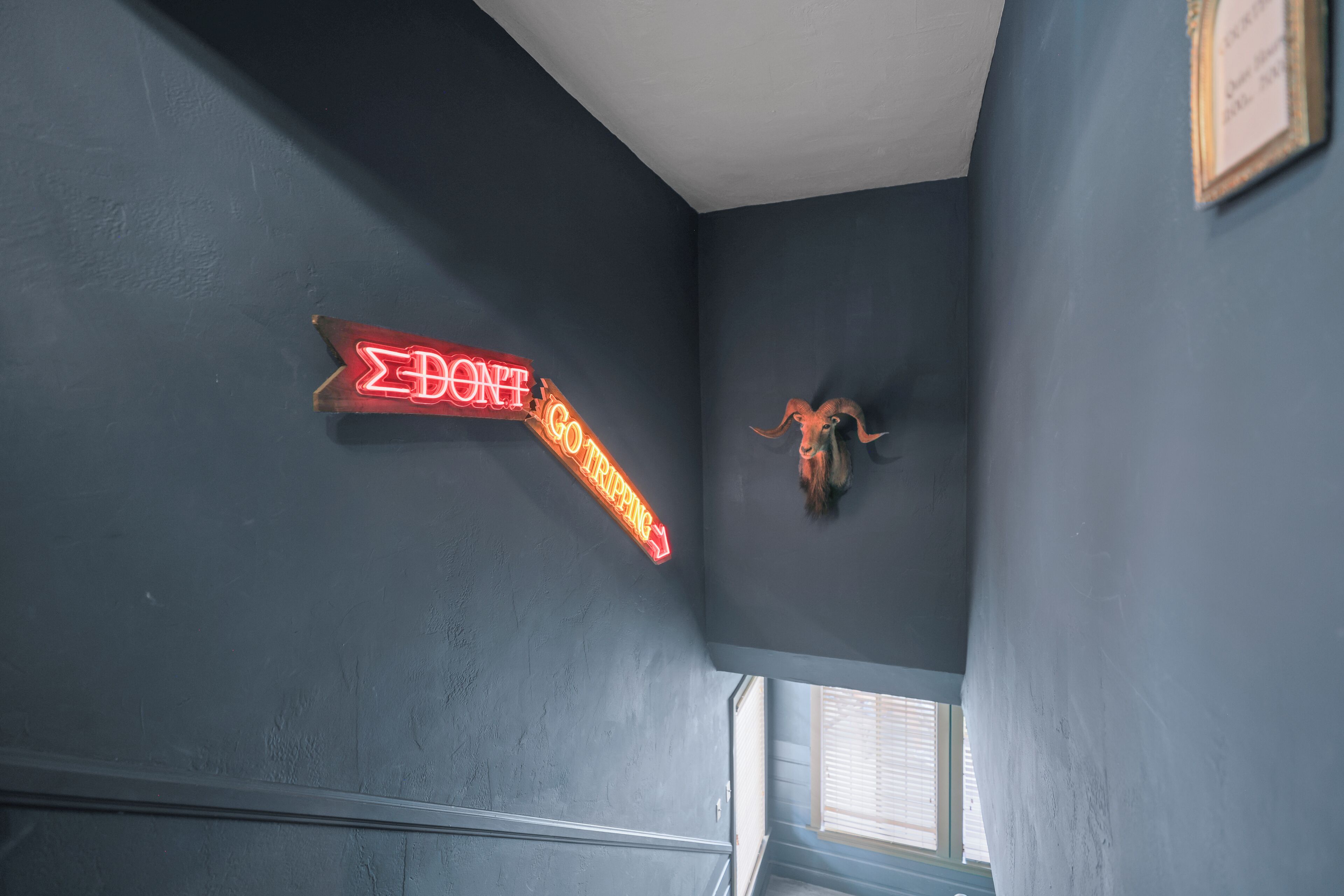 Un letrero de neón que dice "DON'T TRIP" en un pasillo con pared texturizada y un cuadro enmarcado.