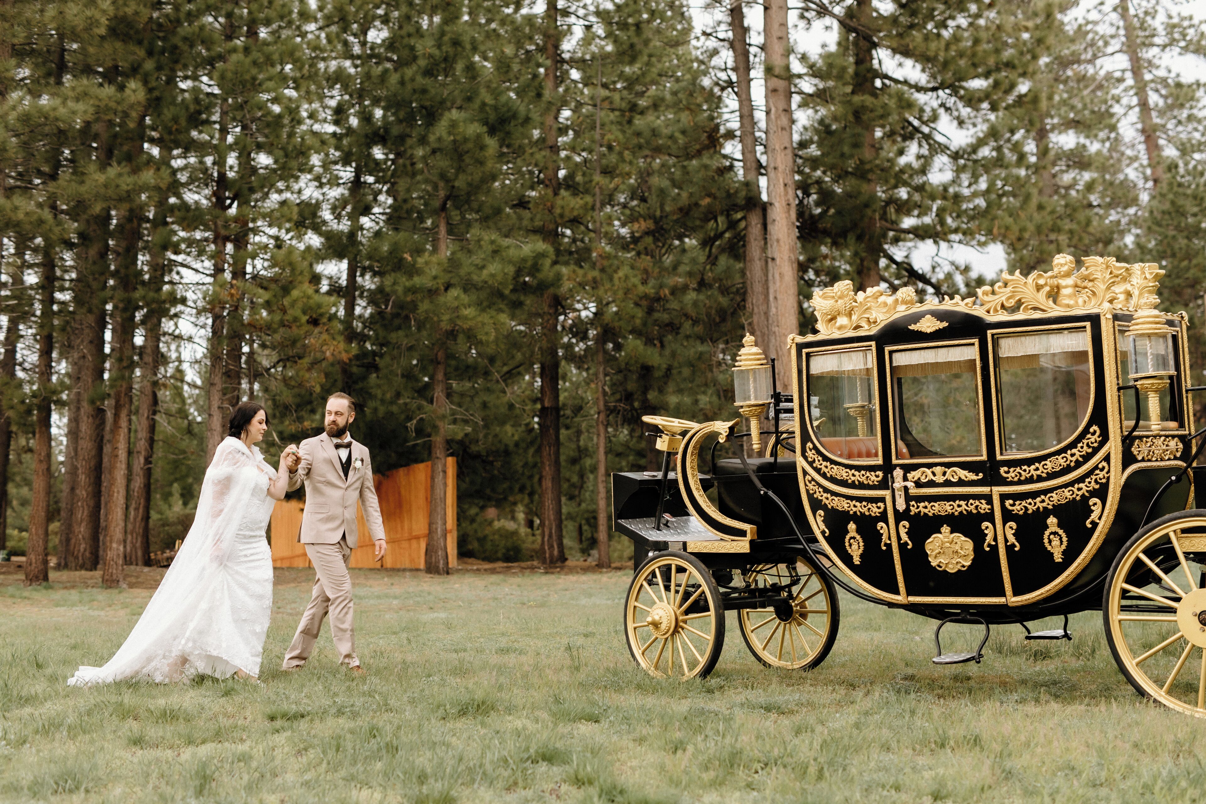 A couple walking beside a black and gold carriage in a grassy field with tall trees in the background.