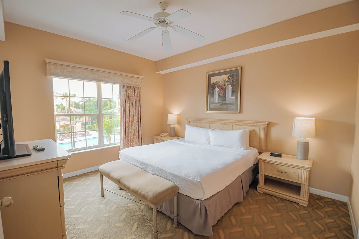 A hotel room with a bed, bedside tables, a desk, a chair, a window with curtains, a ceiling fan, and a painting on the wall.