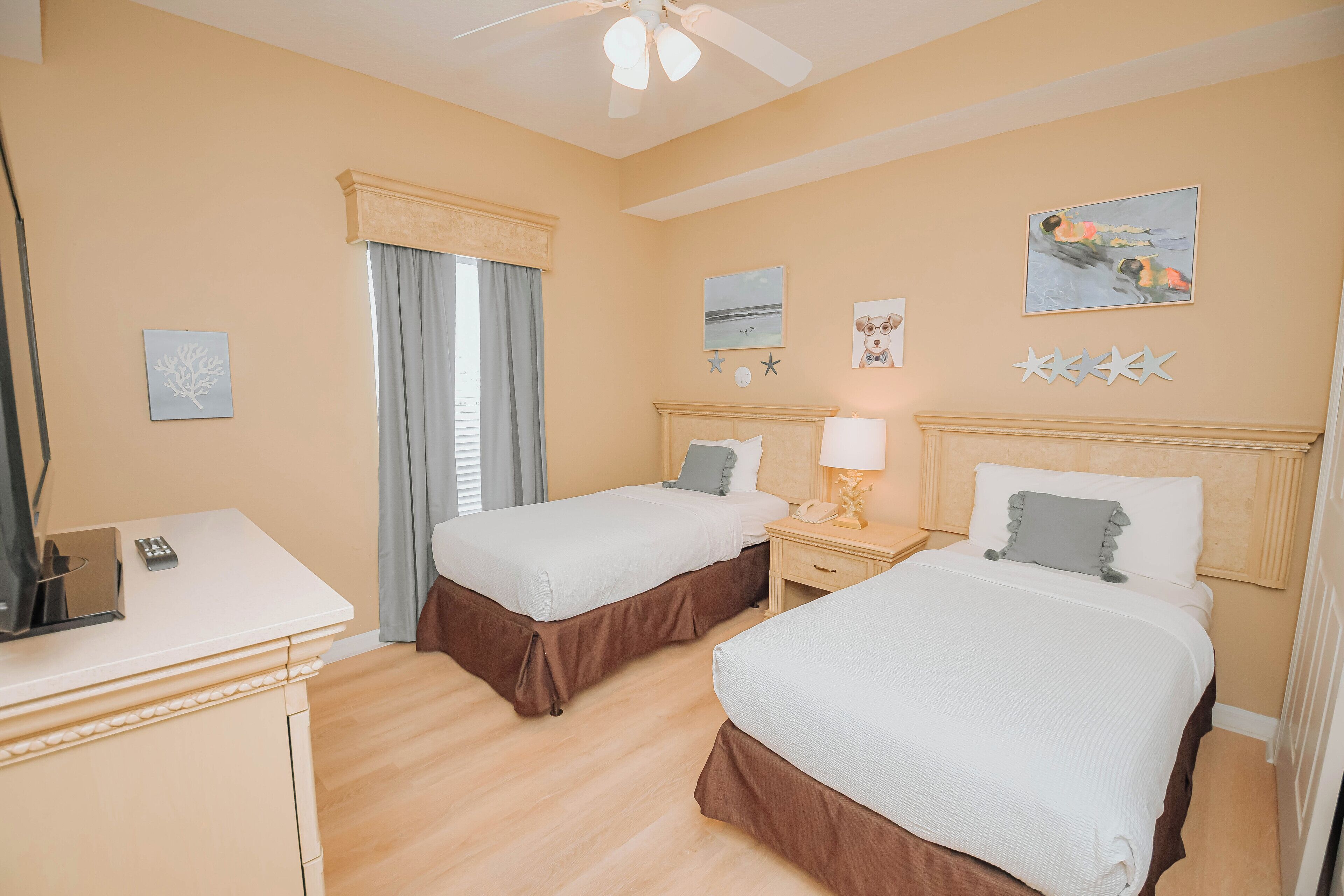 A hotel room with two beds, a desk, a chair, a ceiling fan, and artwork on the walls.