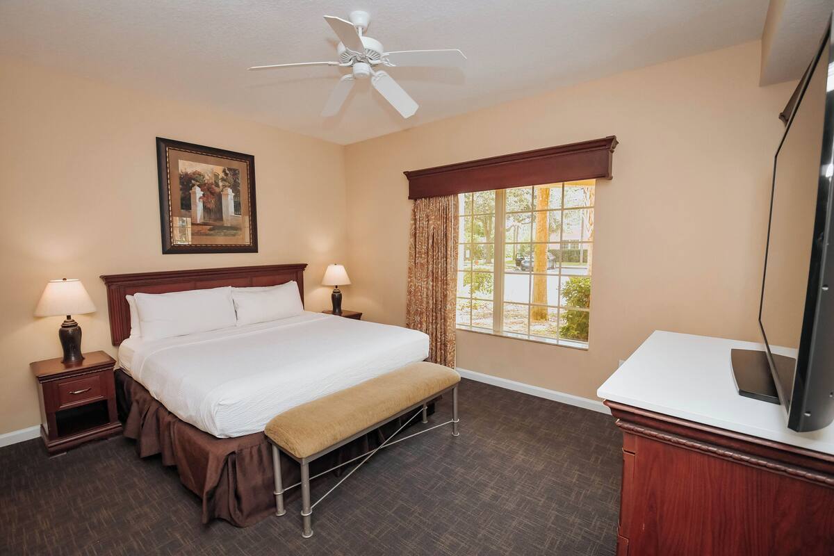 A hotel room with a bed, bedside lamps, a ceiling fan, a window with curtains, and a television on a stand.