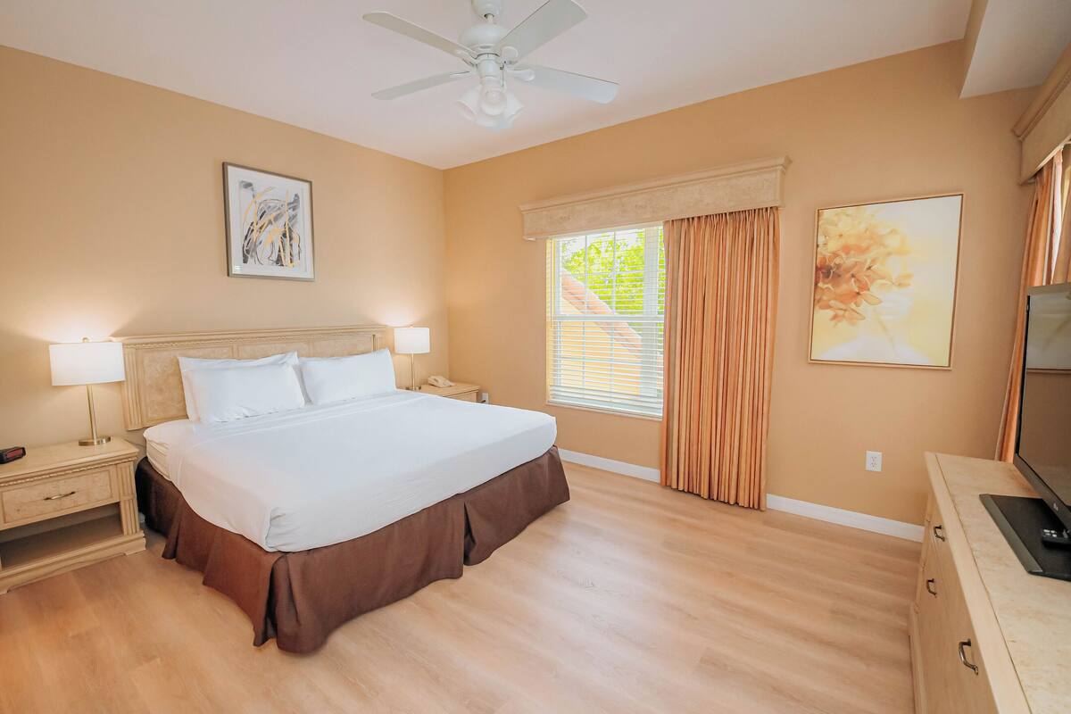 A hotel room with a large bed, bedside lamps, a ceiling fan, a window with curtains, and artwork on the walls.