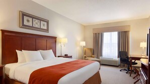 Quarto de hotel com uma cama grande, uma mesa, uma cadeira, uma luminária, um porta-retrato e uma janela com cortinas.