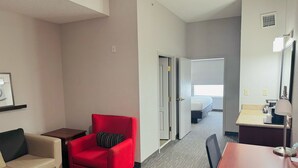 Quarto de hotel com poltrona vermelha, sofá bege, escrivaninha de madeira e cama com vista para o exterior.