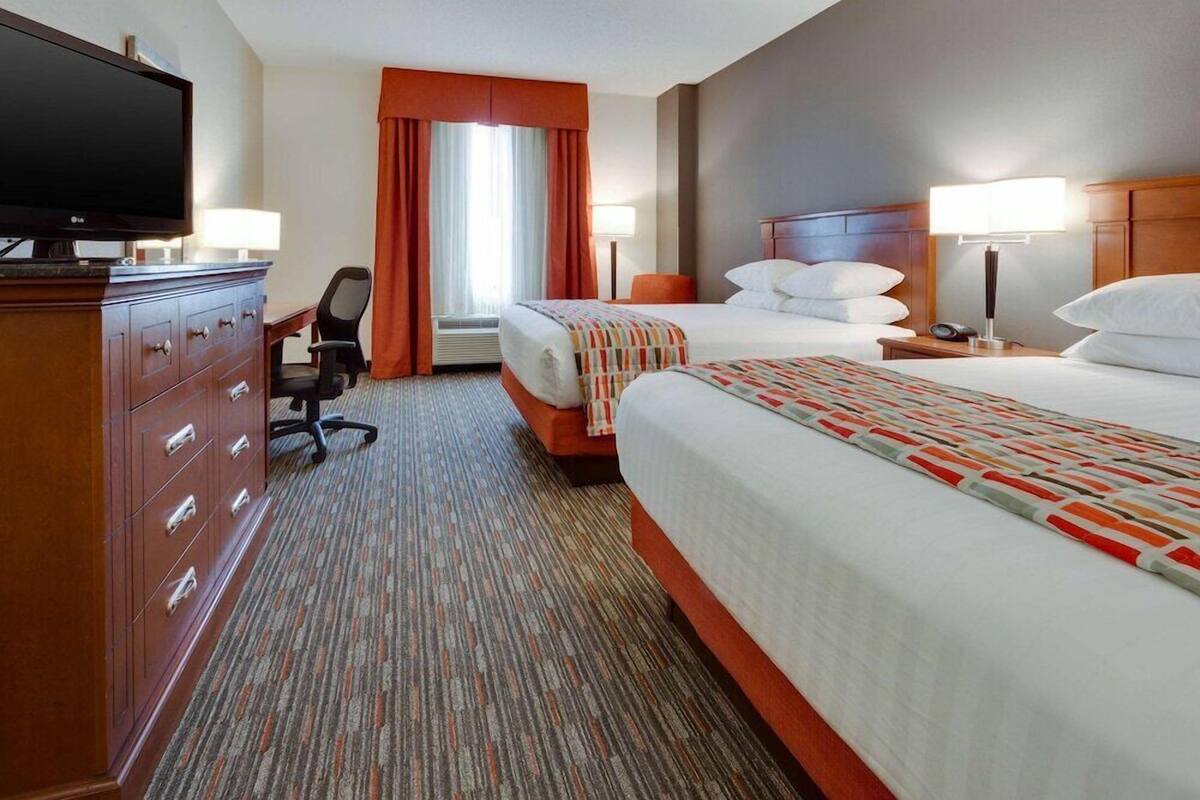 Une chambre d’hôtel équipée d’une télévision, d’un bureau avec une chaise, de deux lits avec des oreillers, d’une lampe et d’une fenêtre avec des rideaux.