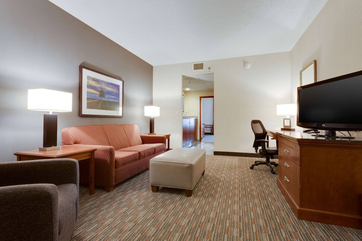 Une chambre d’hôtel équipée d’un canap é, d’un pouf, d’une télévision, d’un bureau et de lampes.