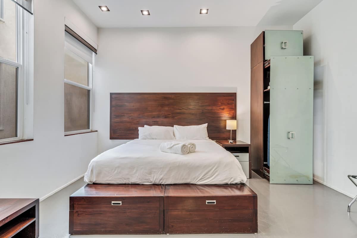 A modern hotel room with a wooden headboard, a bed with white linens, a nightstand, a green cabinet, and a window with blinds.