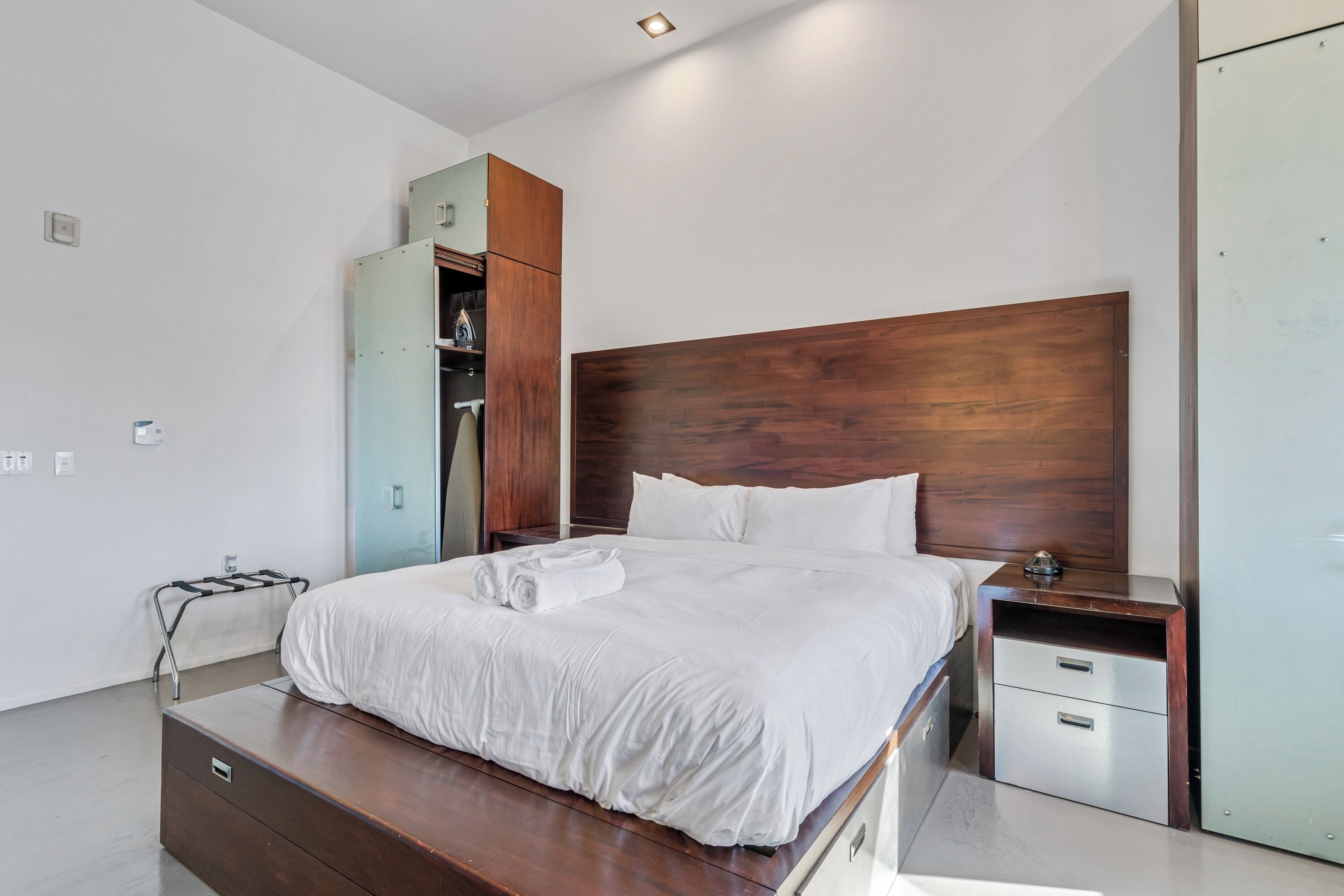 Een moderne slaapkamer met een houten hoofdbord, een groot bed met witte lakens, een nachtkastje, een kledingkast en een inbouwkast.
