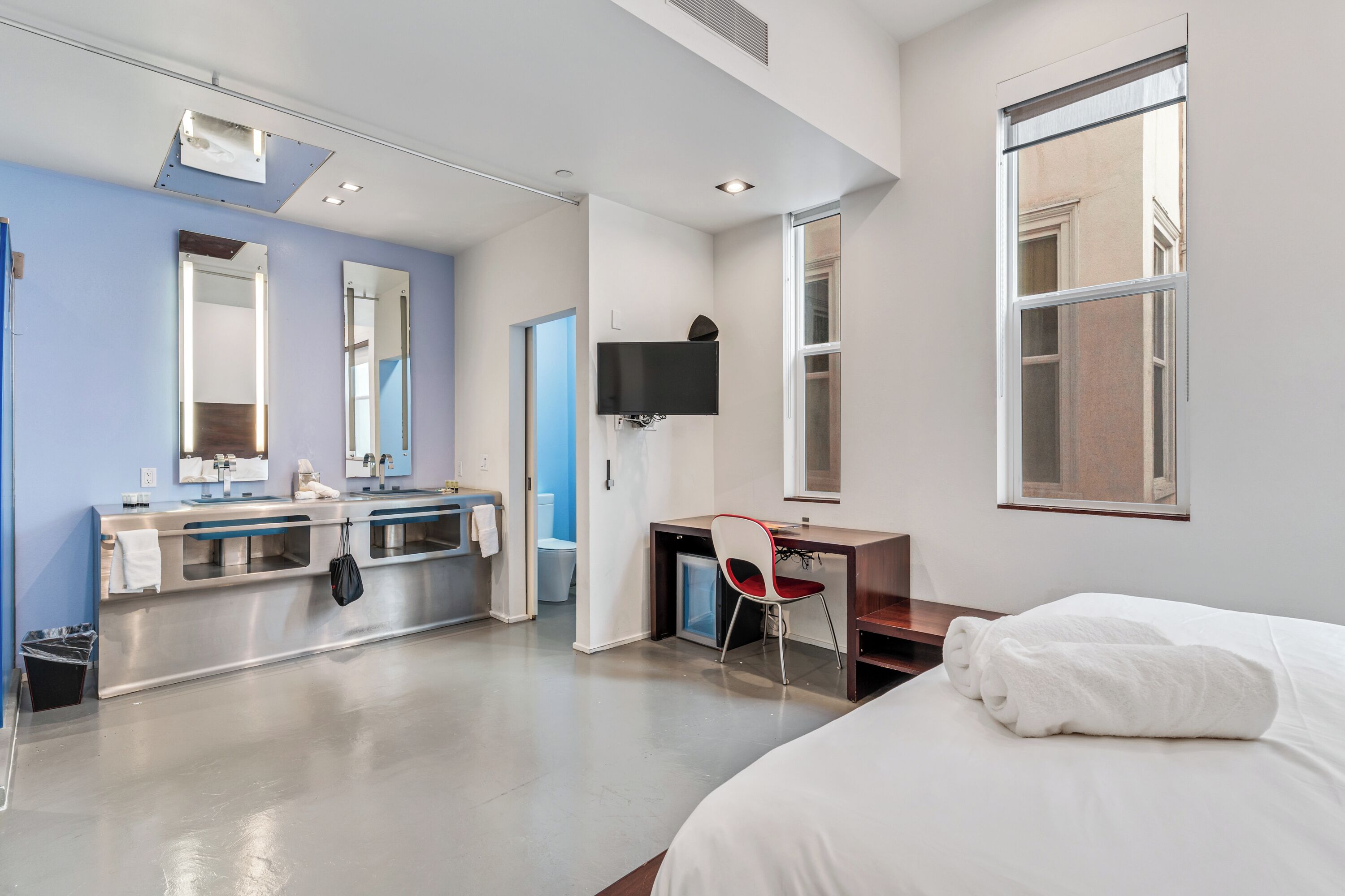 Una camera d'albergo moderna con un letto grande, una scrivania, una sedia, una TV, un bagno con lavandino e specchio, e un'ampia finestra.