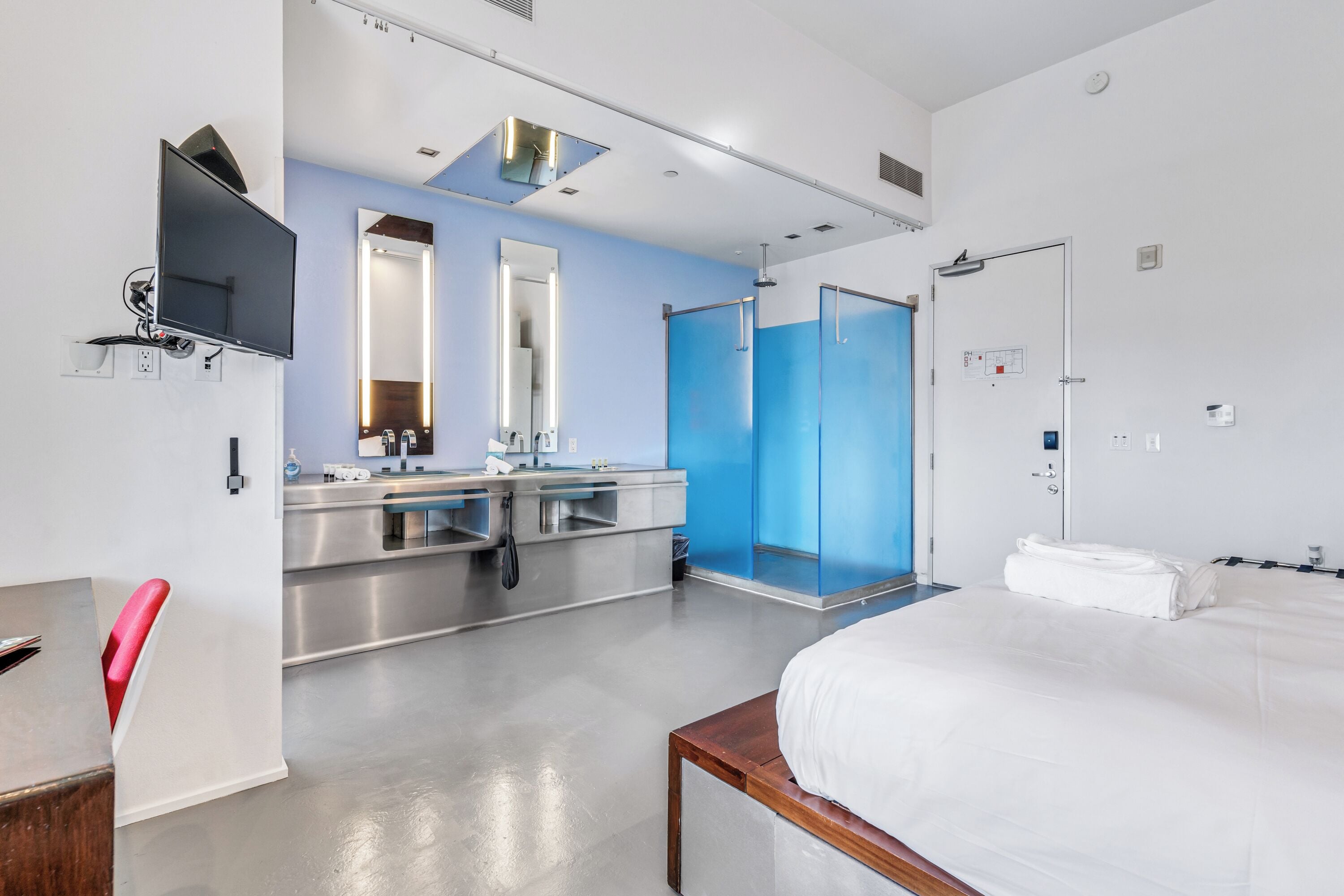 Habitación de hotel con cama, escritorio, silla, baño con ducha y televisor montado en la pared.