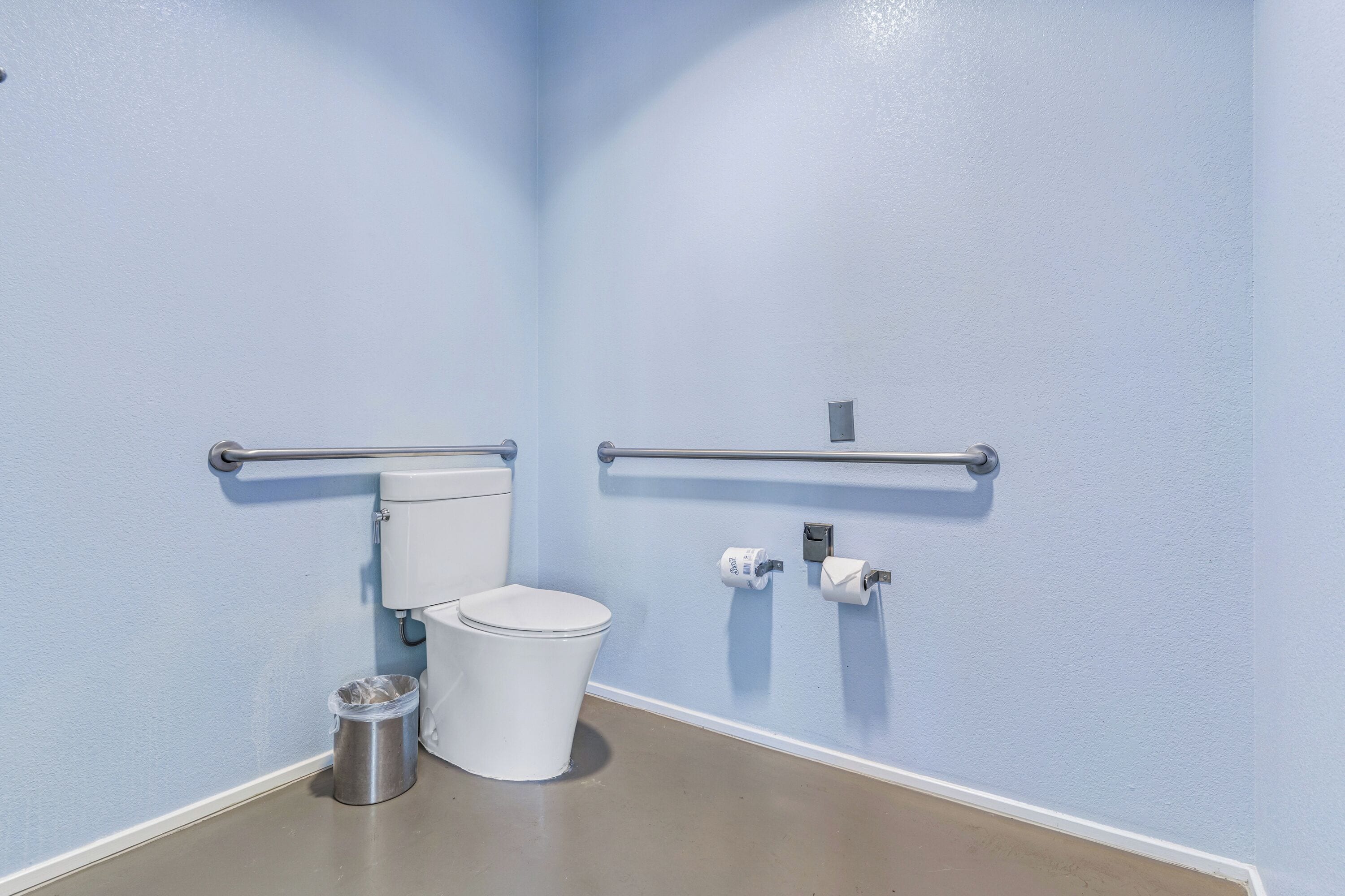 A bathroom with a toilet, a trash can, and two handrails on the walls.