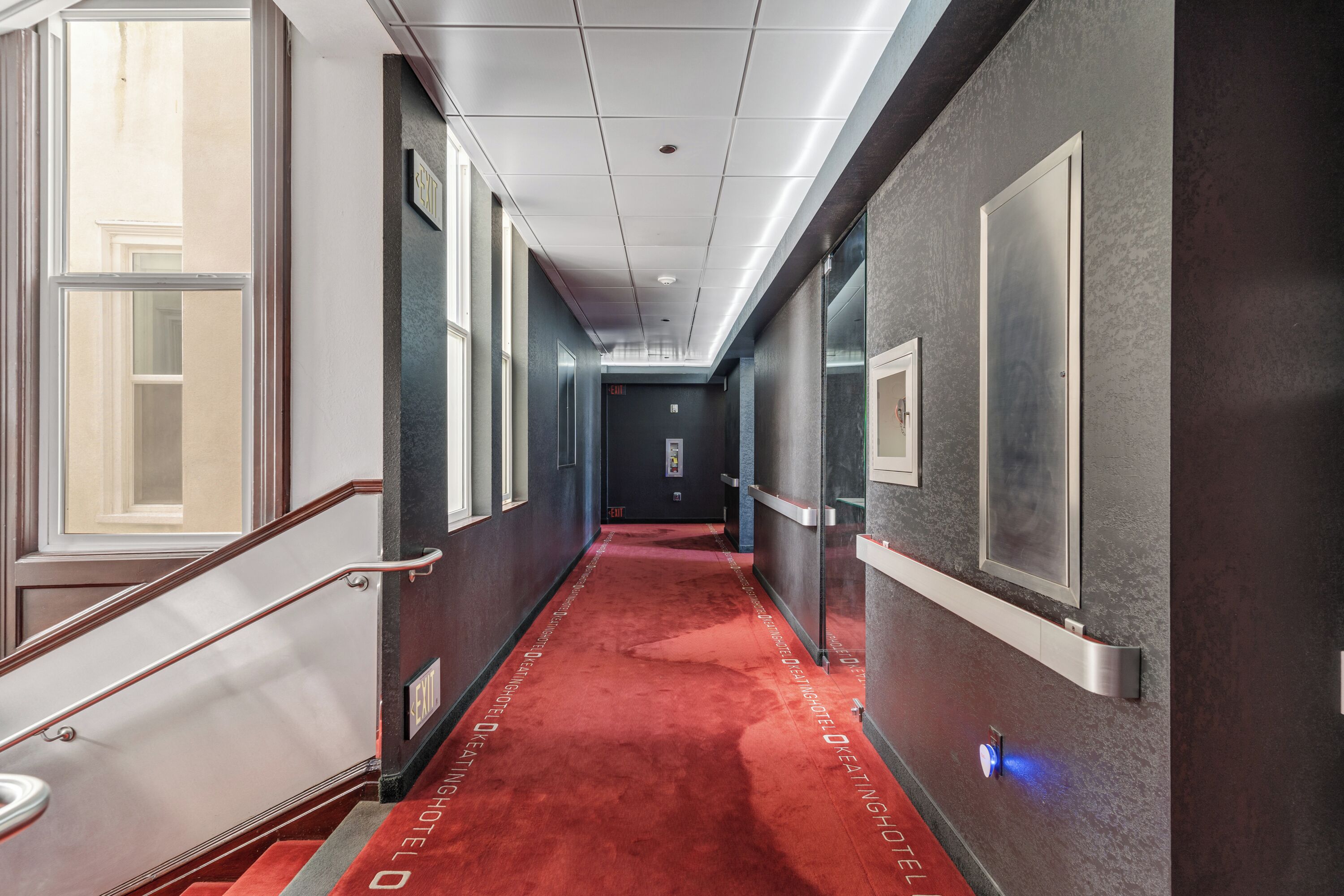 A hotel hallway with a red carpet, a staircase, and a door at the end.