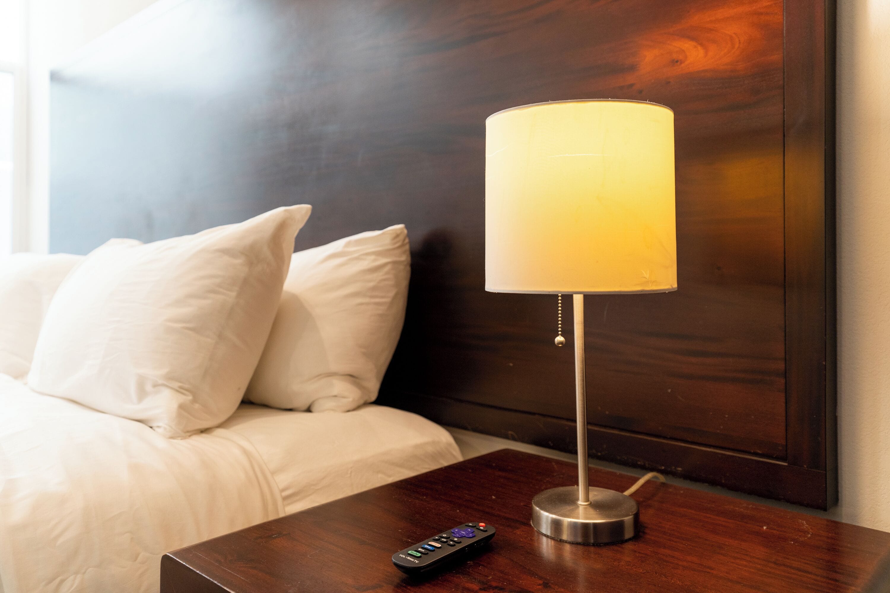 A bed with white bedding, a wooden headboard, a bedside table with a lamp, and a remote control.