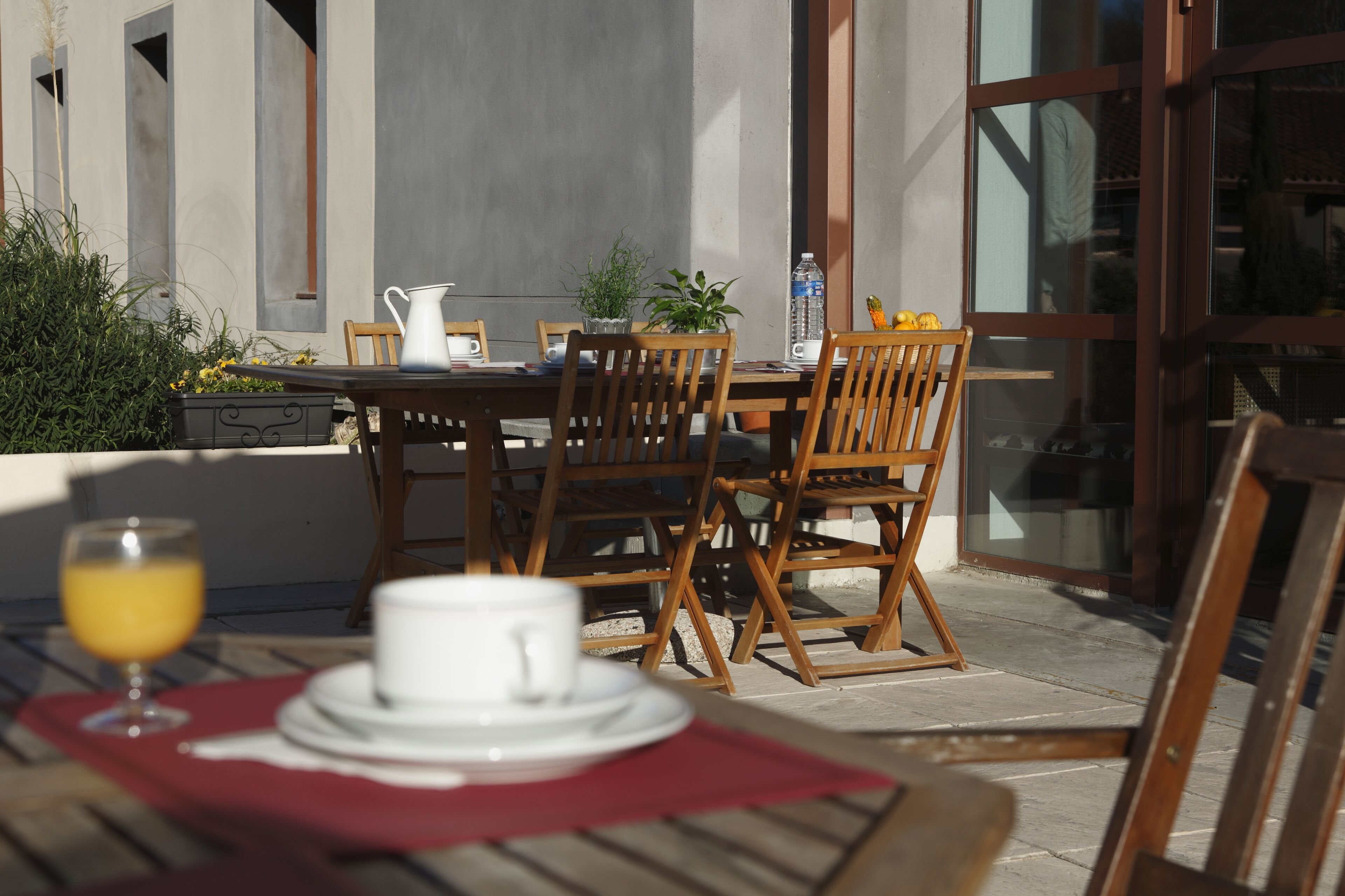 An outdoor dining area with wooden chairs and a table, a pitcher, a glass of orange juice, and a white cup on a red napkin.