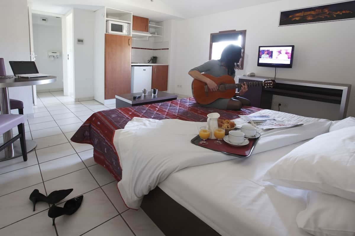 A hotel room with a bed, a dining table, a laptop, a television, and a guitar.