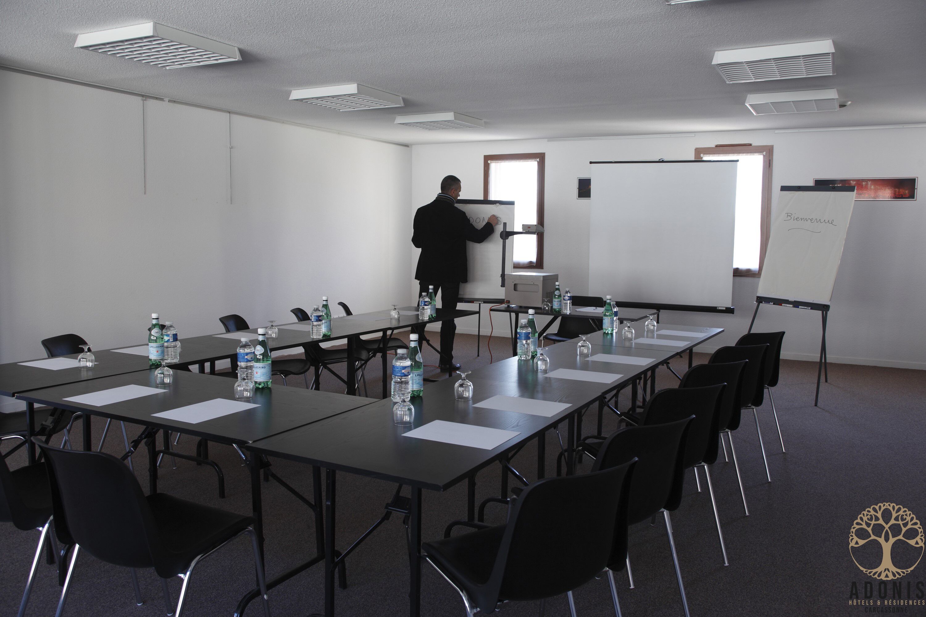 Una sala de conferencias con una pizarra, una persona de pie junto a ella, y un letrero en la pared que dice 'Bienvenue'.