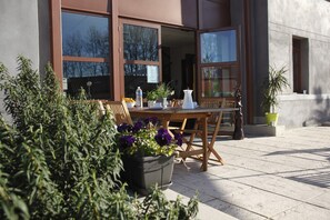 Un patio al aire libre con una mesa y sillas de madera, plantas en macetas y una vista despejada del interior del edificio a través de amplias ventanas.