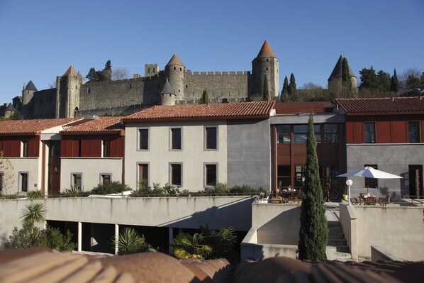 Un hotel con un patio que presenta un castillo al fondo, un edificio moderno con terraza y un asta de bandera.