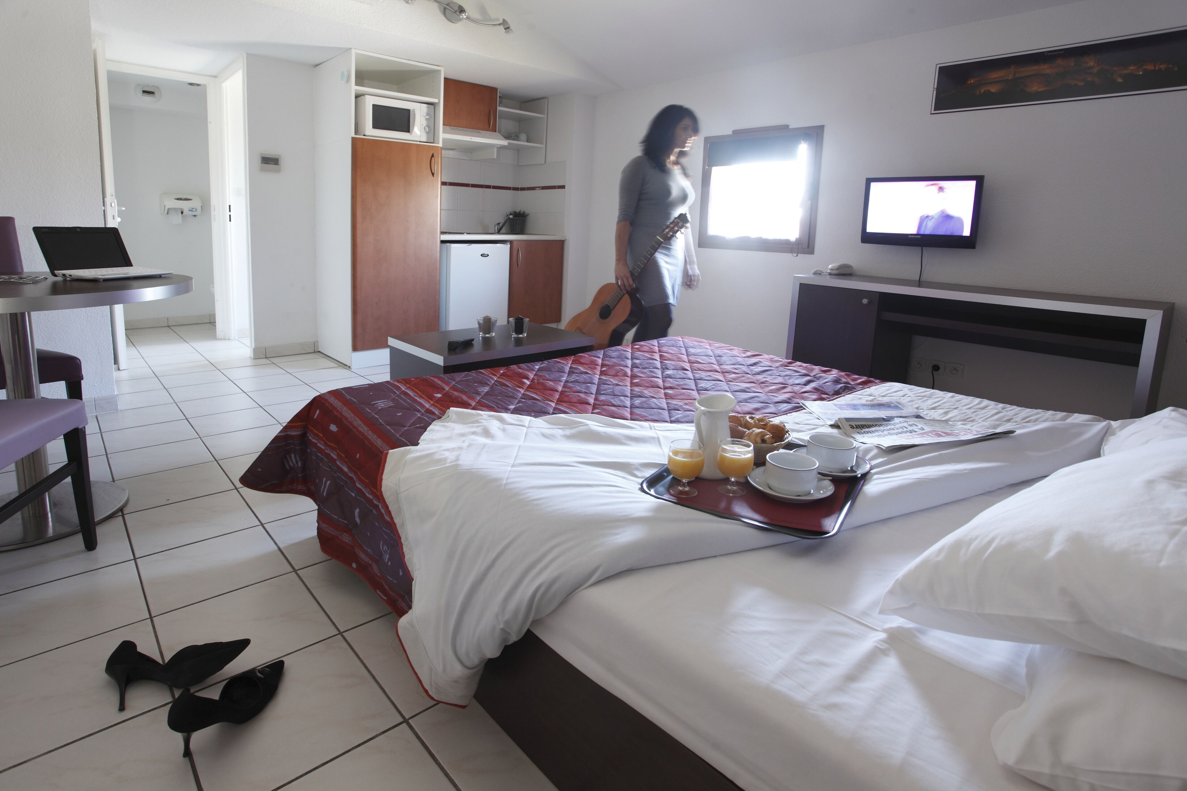 A hotel room with a bed, a desk with a laptop, a TV, and a kitchenette.