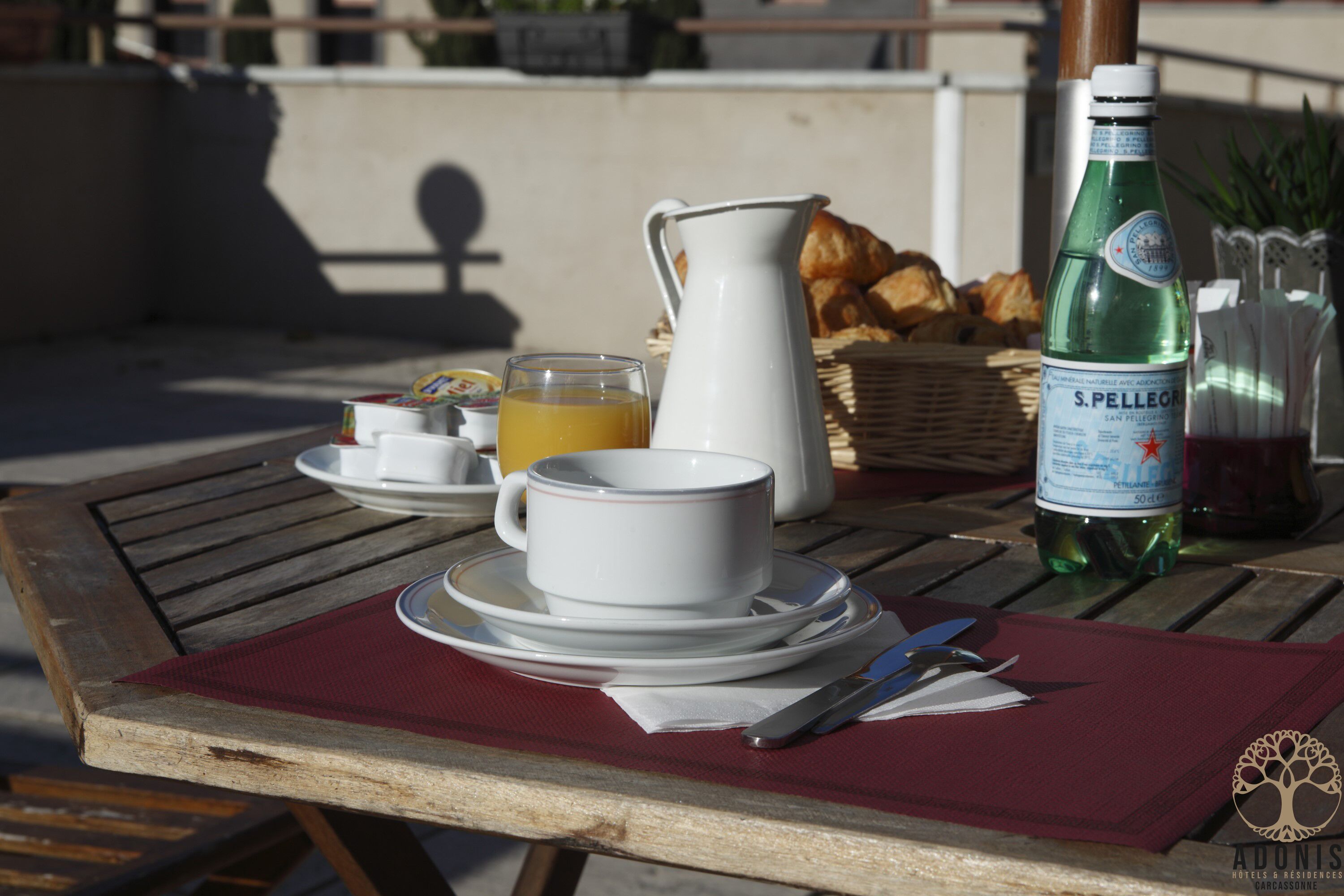 Une table dressée avec une carafe, des tasses et une bouteille d’eau S. Pellegrino, ainsi que des croissants et un panier de pain.