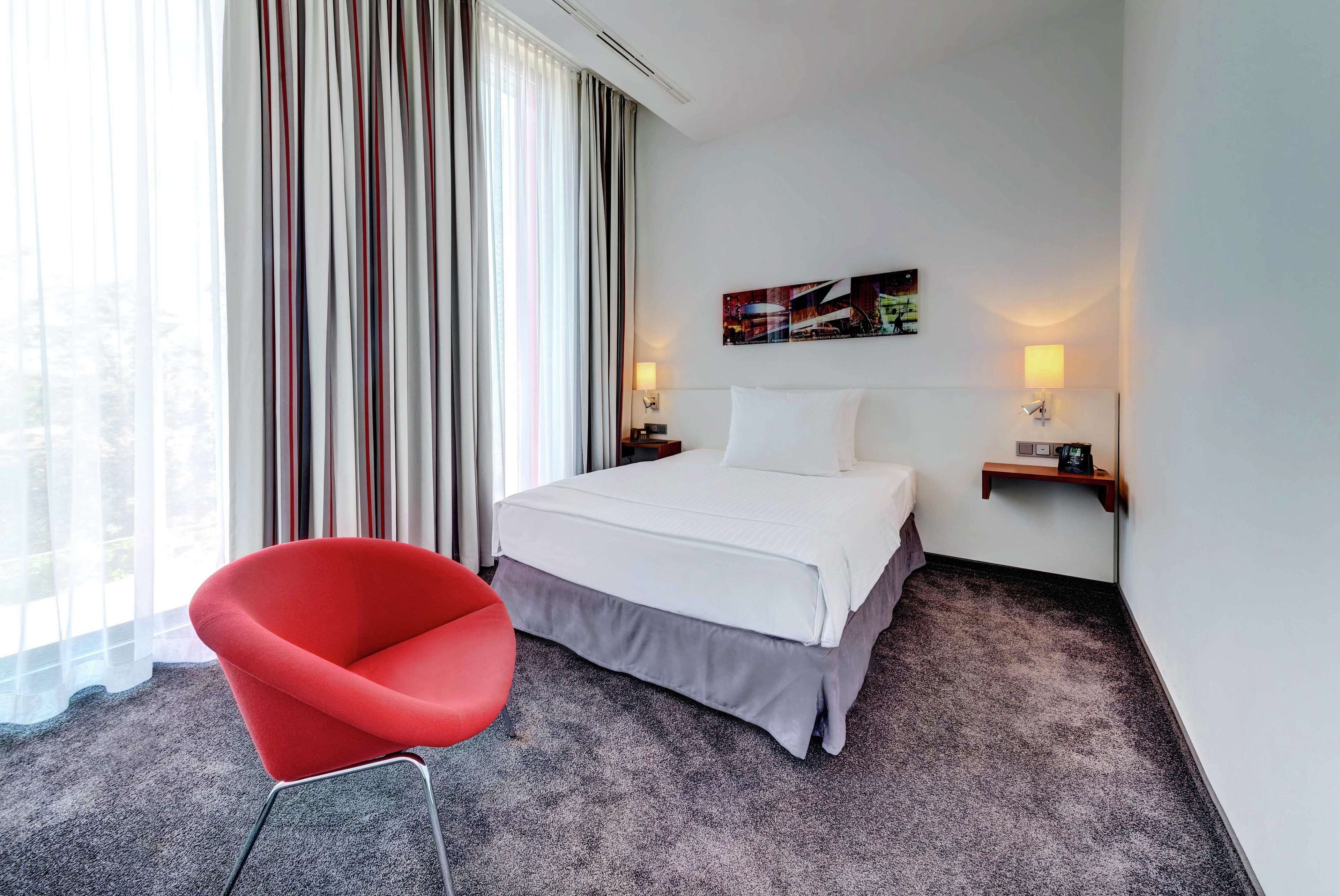 A hotel room with a bed, a red chair, a nightstand with a lamp, and a painting on the wall.