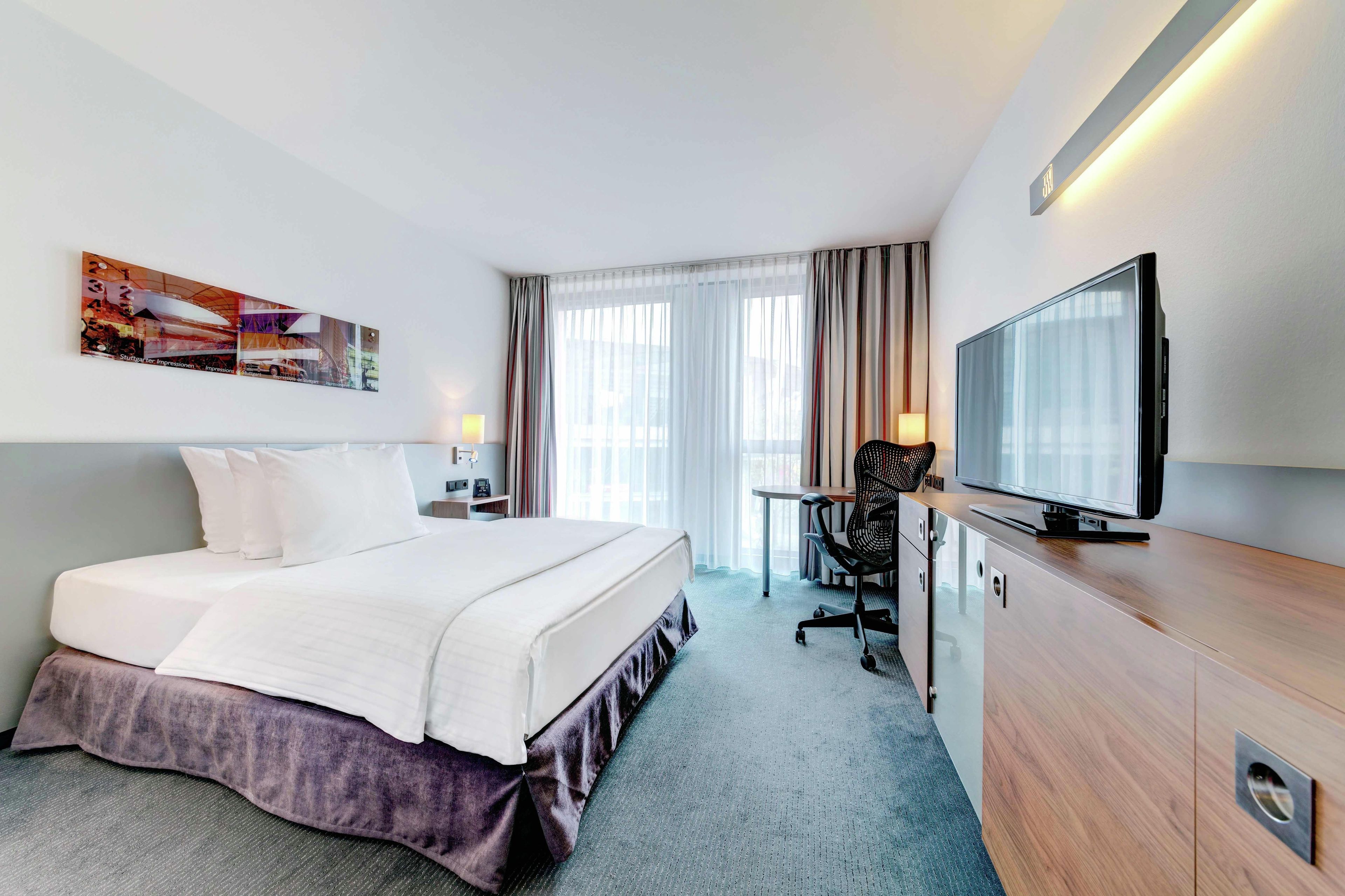 A hotel room with a bed, a desk, a chair, a television, and a large window with curtains.