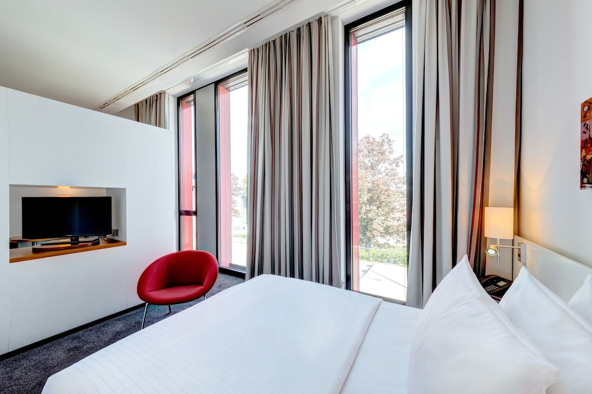 A hotel room with a large bed, a flat-screen TV, a red chair, and a window with curtains.