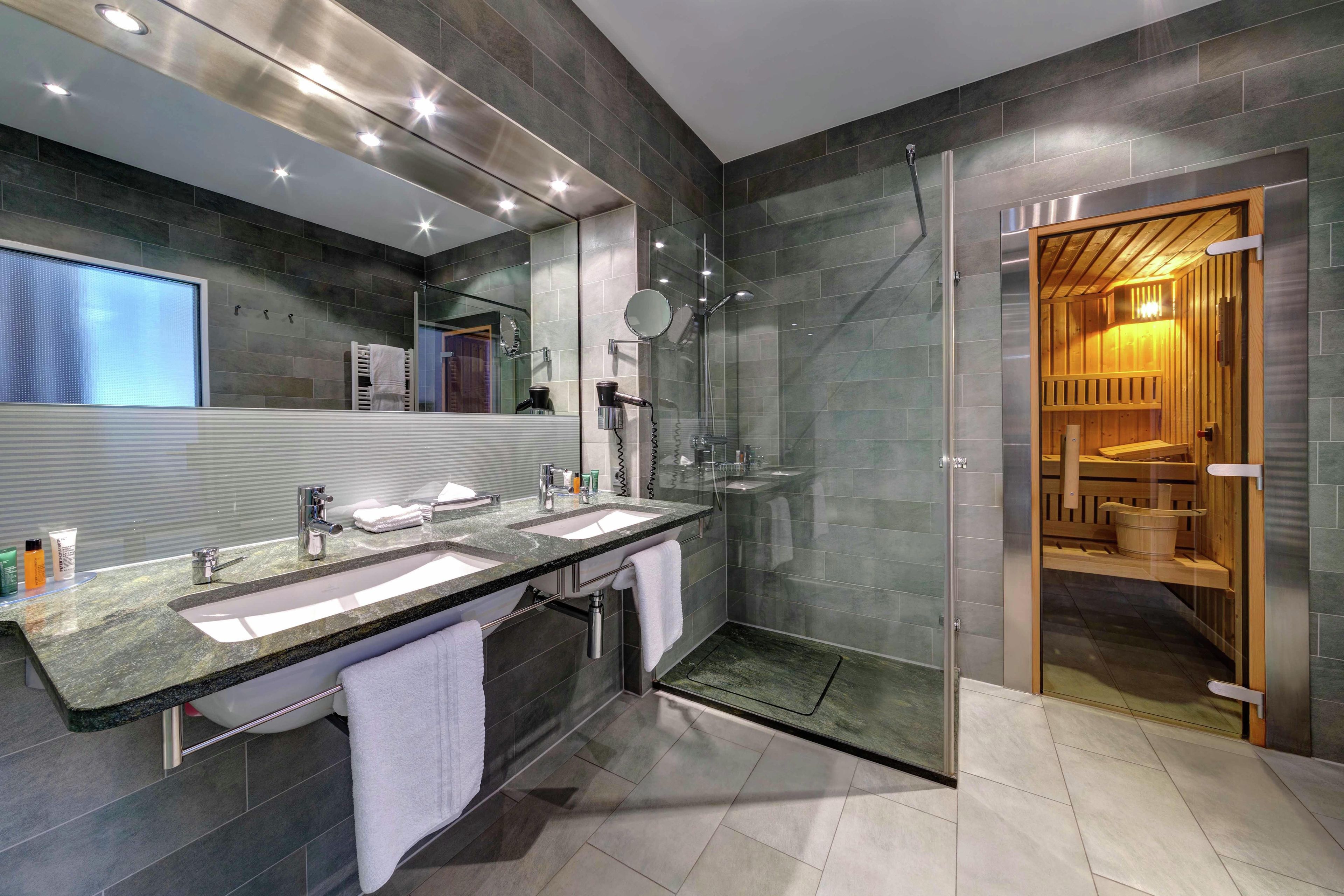Un baño moderno con dos lavabos, cabina de ducha de vidrio y sauna visible a través de una puerta abierta.