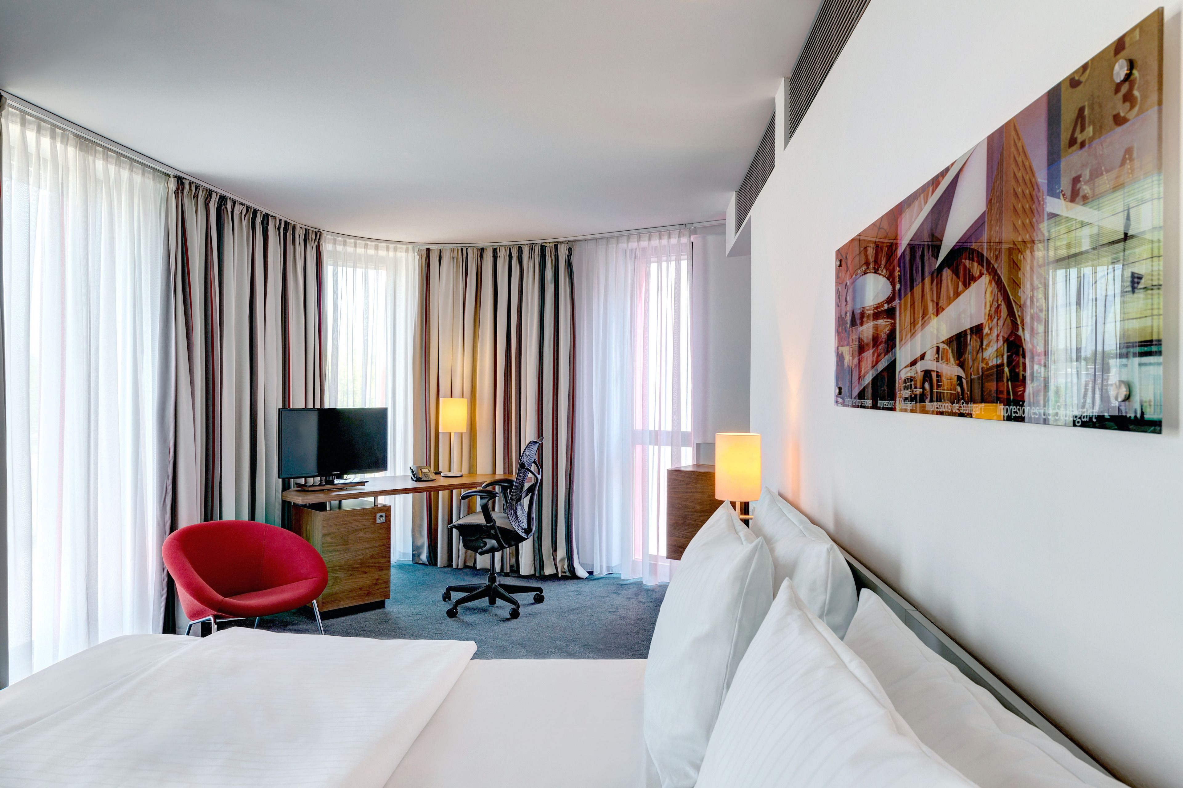 Quarto de hotel com uma cama, uma mesa com cadeira, televisão e uma janela grande com cortinas.